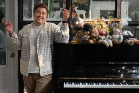 พี่โมผู้ก่อตั้งสตูดิโอ ยืนยิ้มหลับตาในเสื้อแจ็กเก็ตถักสีครีมเทา ข้างเปียโนที่วางตุ๊กตาและดอกไม้