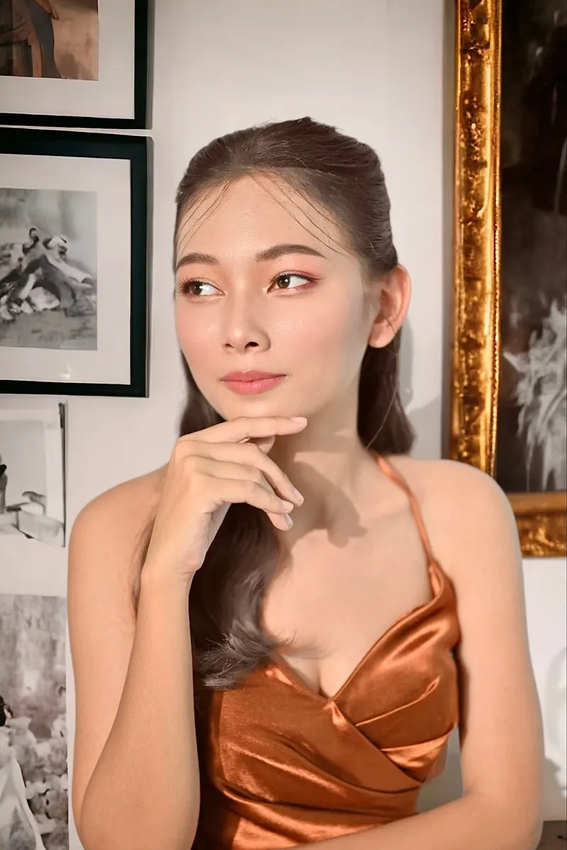 แต่งหน้าออกงานลุคหวานนู้ด ผมรวบครึ่งหัวปล่อยยาว สวมชุดผ้าซาตินสีน้ำตาลส้มคล้องคอ ยกมือแตะคาง มองไปด้านข้าง ฉากกรอบรูปทอง