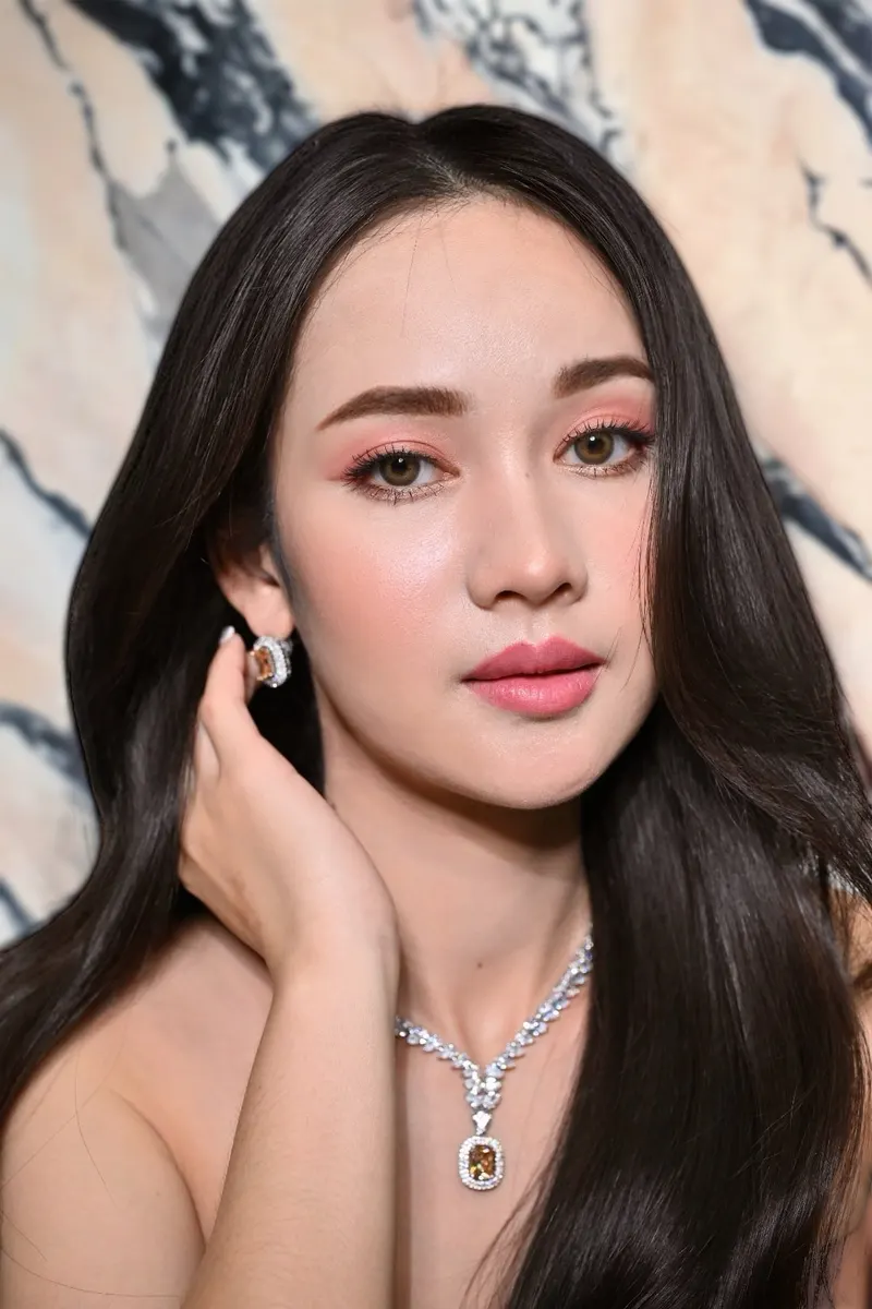 ผมดำยาวตรงสลวย ต่างหูและสร้อยเพชรอัญมณีสีส้ม ยกมือแตะหู แต่งตาโทนชมพูพีช ลิปสีชมพูเข้ม ฉากหินอ่อนลายเทา