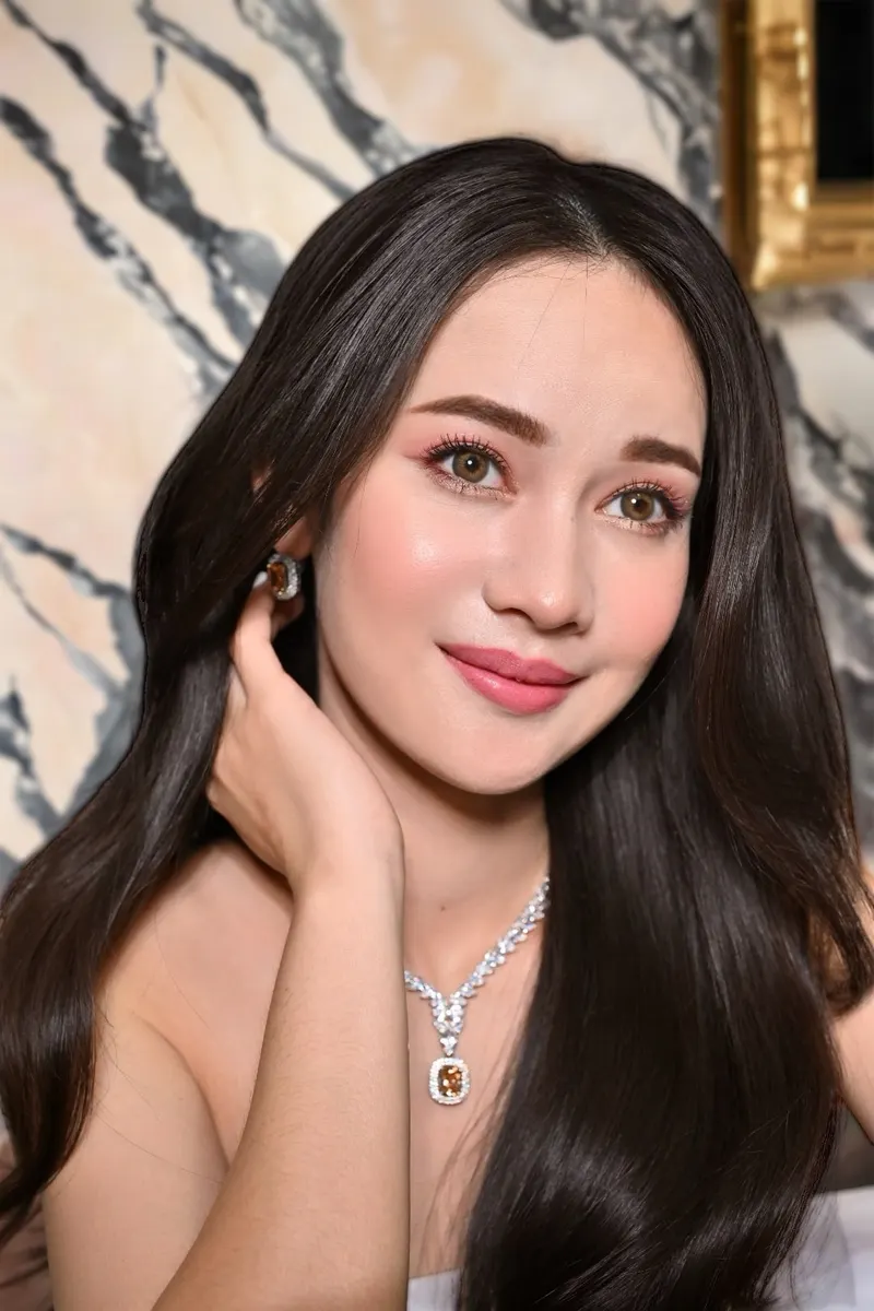 ผมดำตรงยาวปล่อยหน้าอก ยกมือแตะใบหู สร้อยเพชรจี้อัญมณีสีส้ม ปัดแก้มชมพูเด่น ลิปสีโอลด์โรส ฉากหินอ่อนลายเทา