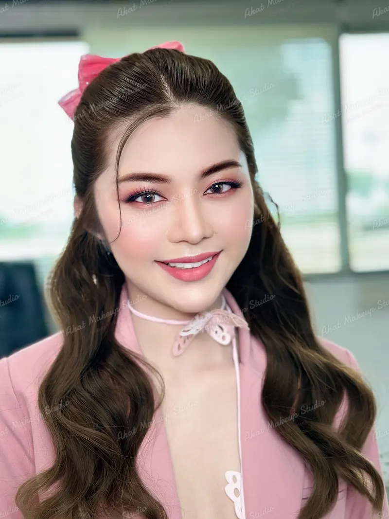 ผมลอนยาวรวบครึ่งหัวผูกโบว์ชมพู สวมแจ็กเก็ตสีชมพูกับโบว์ลูกไม้ที่คอ แต่งตาโทนชมพูม่วง ลิปสีชมพูกลอส ฉากออฟฟิศสว่างเบลอ