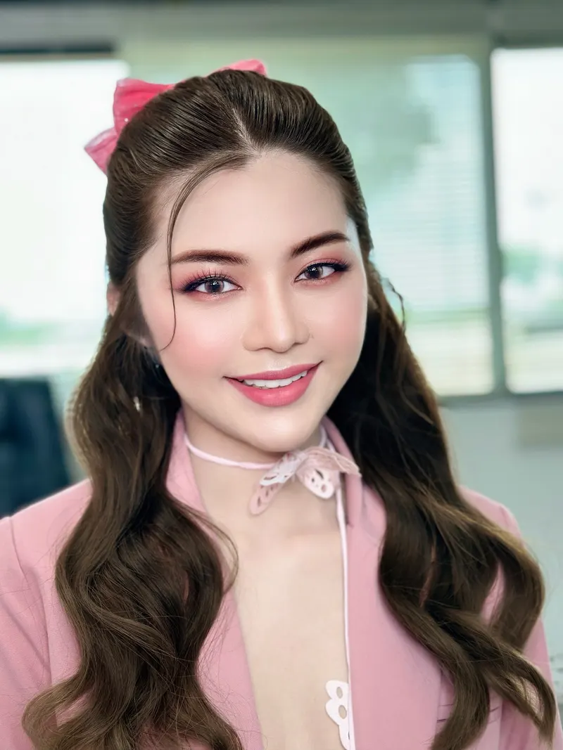 ผมลอนยาวรวบครึ่งหัวผูกโบว์ชมพู สวมแจ็กเก็ตสีชมพูกับโบว์ลูกไม้ที่คอ แต่งตาโทนชมพูม่วง ลิปสีชมพูกลอส ฉากออฟฟิศสว่างเบลอ