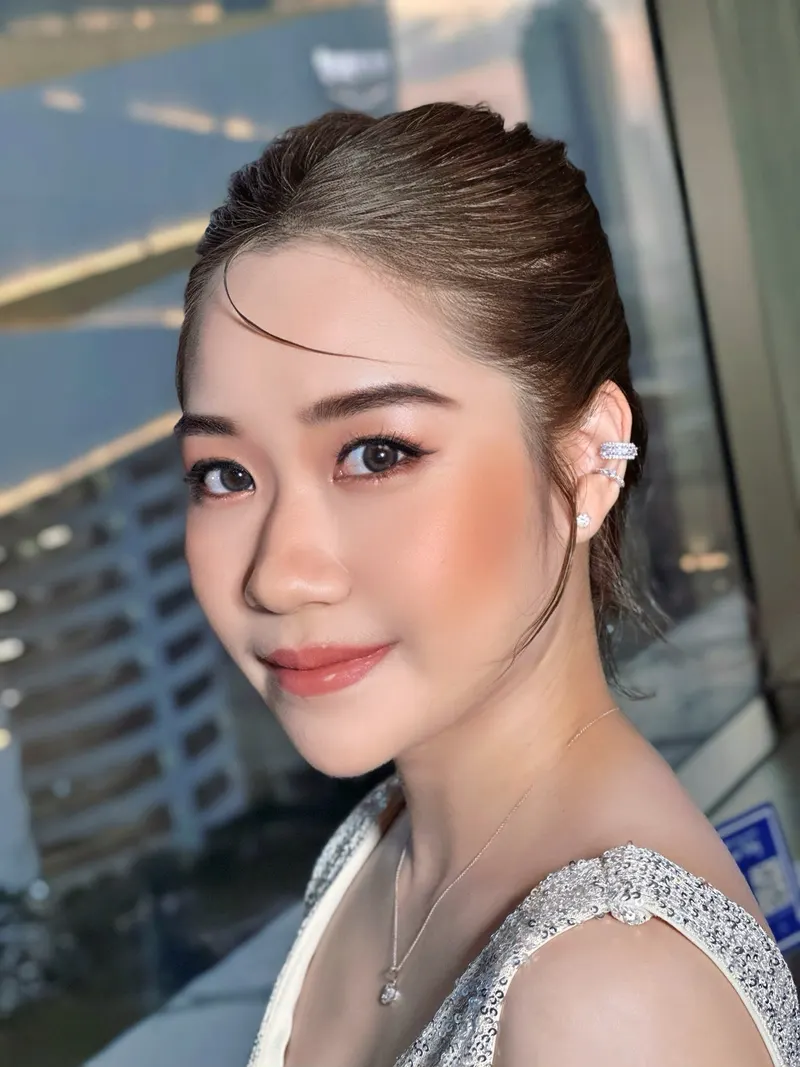 ผมรวบตึงเรียบไปด้านหลัง ต่างหูคัฟที่หู สวมชุดเลื่อมเงินสายเดี่ยว ปัดแก้มพีชส้มเด่น ลิปชมพูส้ม ฉากหลังตึกสูงและกระจก