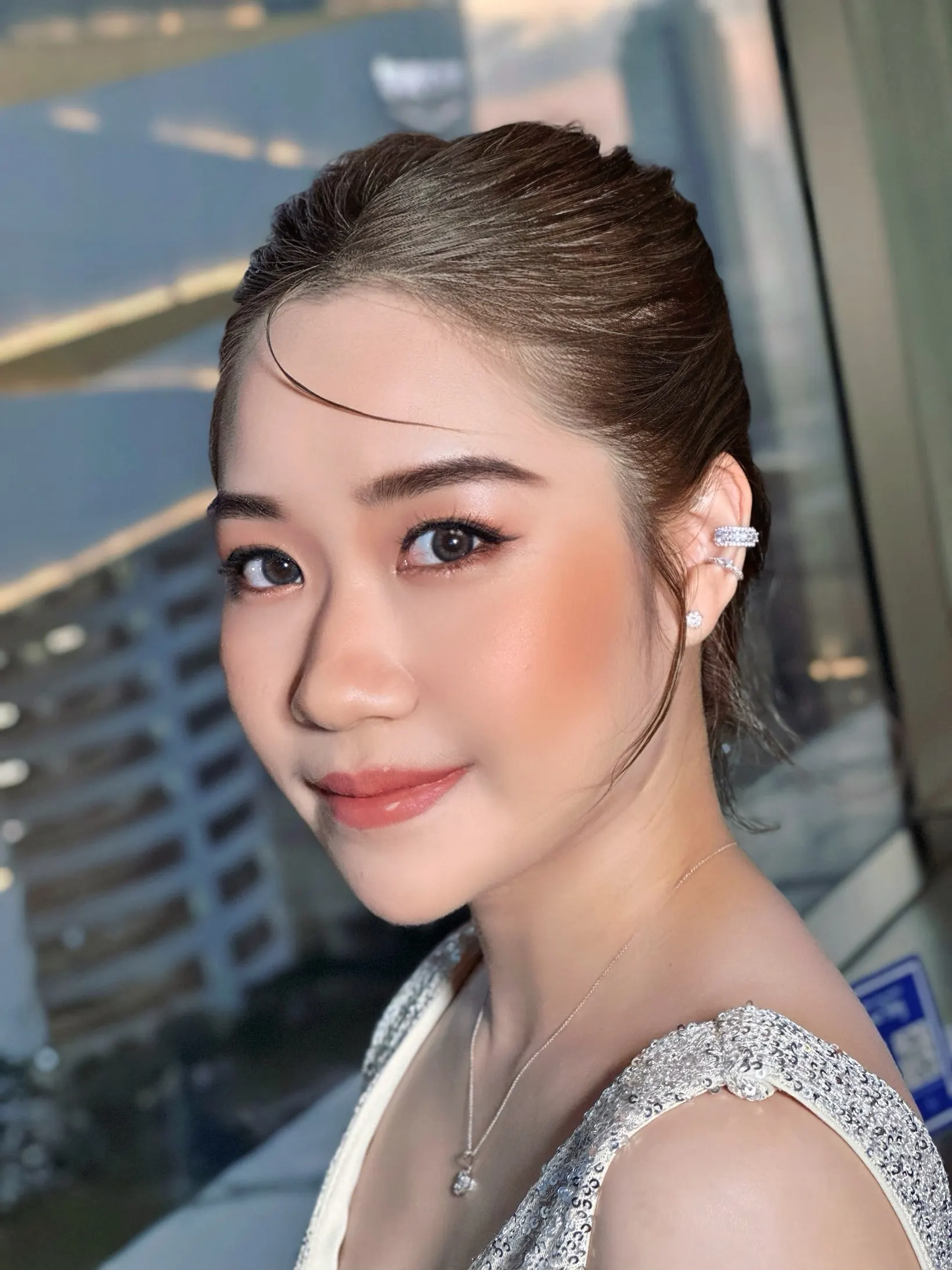 ภาพใกล้ใบหน้าด้านข้างของหญิงสาวผมรวบเรียบไปด้านหลัง ใส่ต่างหูคัฟเพชร ปัดแก้มพีชส้มชัด ลิปกลอสสีพีช ชุดสายเดี่ยวเลื่อมเงิน มีกระจกตึกเป็นฉากหลัง