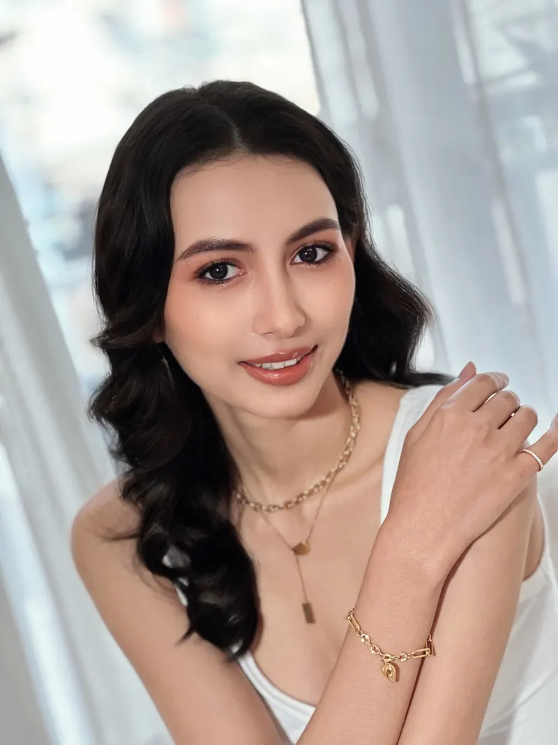 ผมดำลอนใหญ่ปล่อยยาว สวมสร้อยทองหลายเส้นและกำไลโซ่ ยกแขนกอดไหล่ แต่งหน้าโทนนู้ดชมพู ลิปสีพีช ฉากผ้าม่านสีขาว