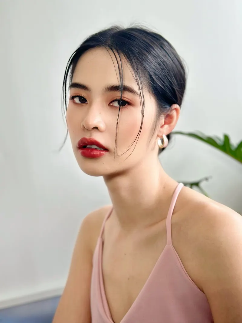 ผมดำรวบต่ำเก็บเรียบปล่อยปอยหน้า สวมต่างหูห่วงทองและเสื้อสายเดี่ยวสีชมพูอ่อน ลิปสีแดงสด แต่งตาโทนส้มแดง สไตล์แฟชั่น