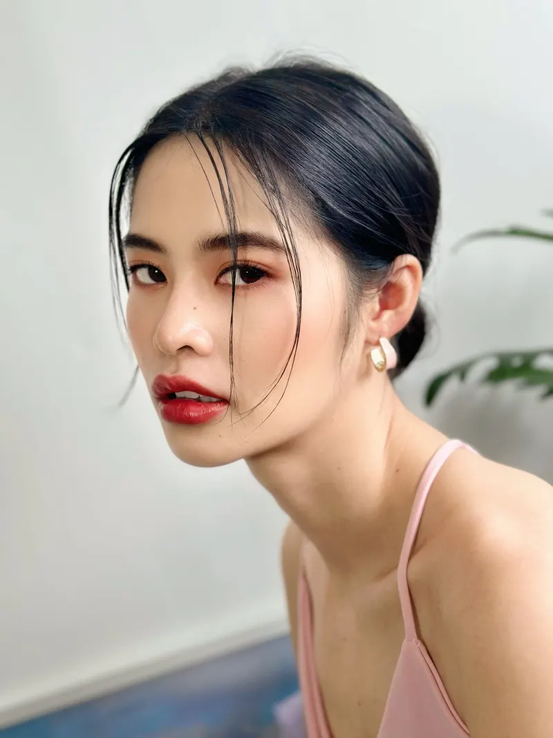 ภาพข้างใบหน้า ผมดำรวบต่ำปล่อยปอยข้างแก้ม ลิปสีแดงสด แต่งตาโทนส้มแดง ต่างหูห่วงทอง ชุดสายเดี่ยวสีชมพูอ่อน ฉากห้องสีขาว