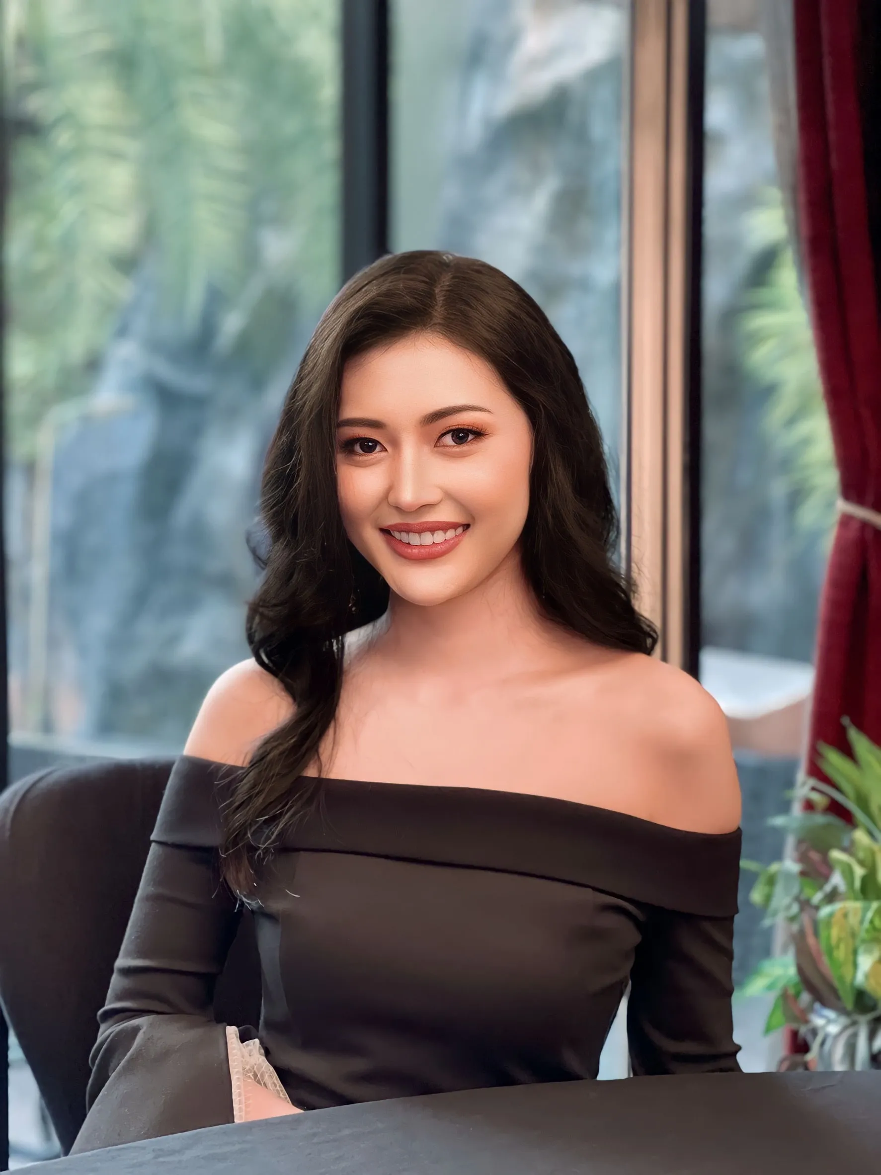 ภาพถ่ายใกล้ของหญิงสาวผมดำลอนใหญ่ปล่อยยาว นั่งยิ้มข้างหน้าต่างบานใหญ่ ใส่เสื้อเปิดไหล่สีดำ ตาโทนส้มพีชคมชัด ลิปสีน้ำตาลส้ม