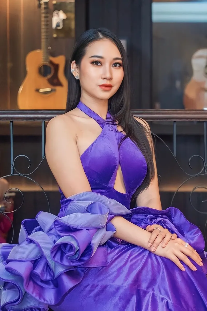 ผมดำยาวตรงนั่งหน้ากีตาร์ สวมชุดกำมะหยี่สีม่วงสดคล้องคอ แต่งหน้าลุคส้มพีชคมชัด ลิปสีส้มแดง ต่างหูตกแต่งใบหูหลายจุด