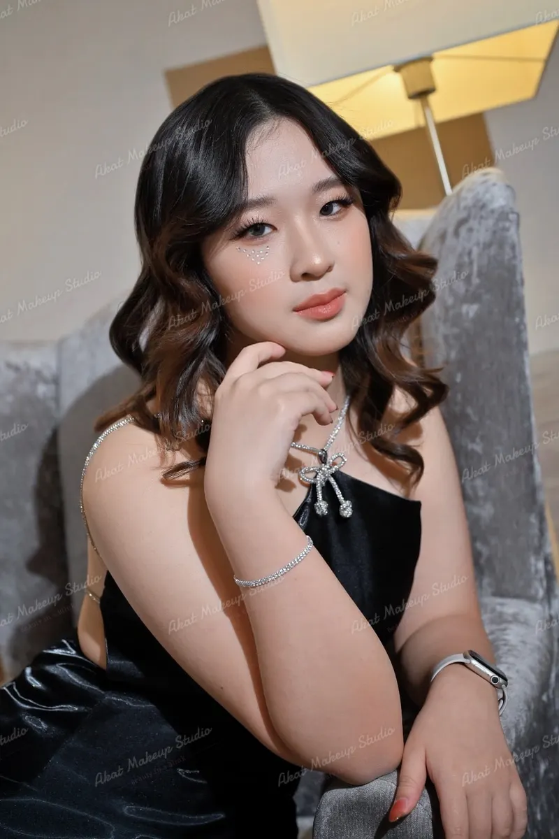 ผมม้าลอนเกลียวสีน้ำตาลเข้มปลายทอง ประดับกลิตเตอร์ใต้ตา ชุดสายเดี่ยวสีดำและสร้อยโบเพชร นั่งบนโซฟาหุ้มกำมะหยี่สีเทา