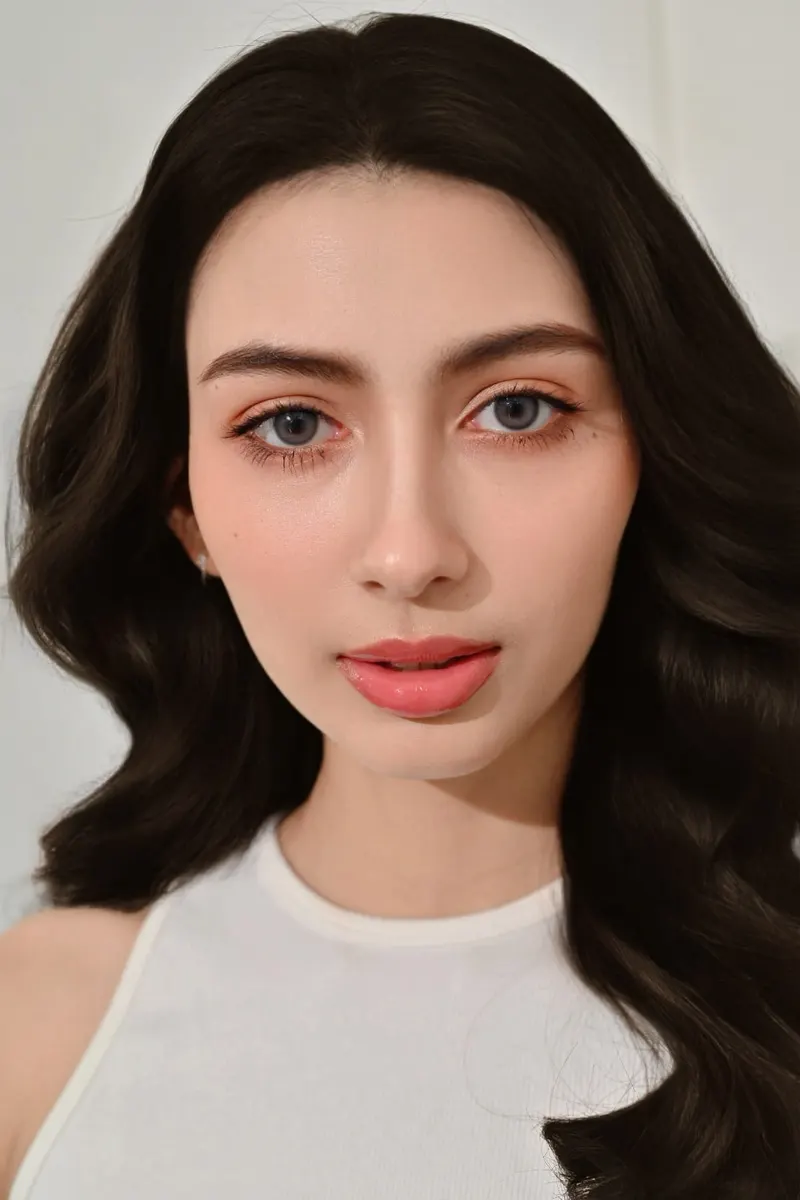 ผมดำลอนใหญ่ปล่อยยาวสองข้าง ชุดแขนกุดสีขาว แต่งตาส้มพีชนุ่ม ขนตางอน ลิปสีส้มคอรัล ผิวเนียนฉ่ำ ฉากสีขาวเรียบ