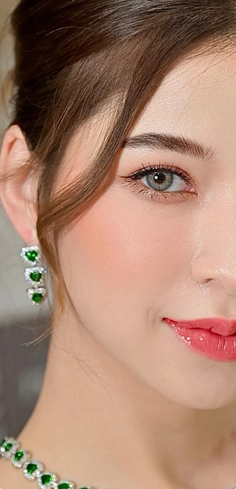 ภาพใกล้ใบหน้าครึ่งซีก ผมน้ำตาลปัดข้างแก้ม ต่างหูและสร้อยมรกตรูปหัวใจ ขนตายาวเส้นไลเนอร์บาง ปัดแก้มชมพูพีช ลิปสีชมพูกลอส