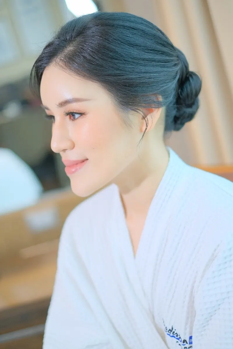 ผมสีดำเทาเกล้ามวยต่ำเก็บเรียบ หันหน้าไปด้านข้างในเสื้อคลุมสีขาว ผิวฉ่ำเรียบ ลิปสีชมพูนู้ด ฉากหลังห้องพักเบลอ