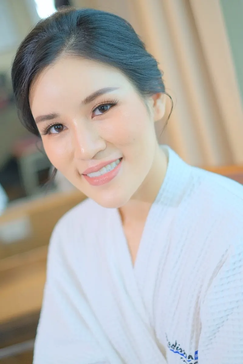 ผมดำเกล้ามวยต่ำ ยิ้มกว้างในเสื้อคลุมสีขาว แต่งตาธรรมชาติ ลิปสีพีชนู้ด ผิวเนียนสว่าง ฉากหลังห้องพักเบลอ