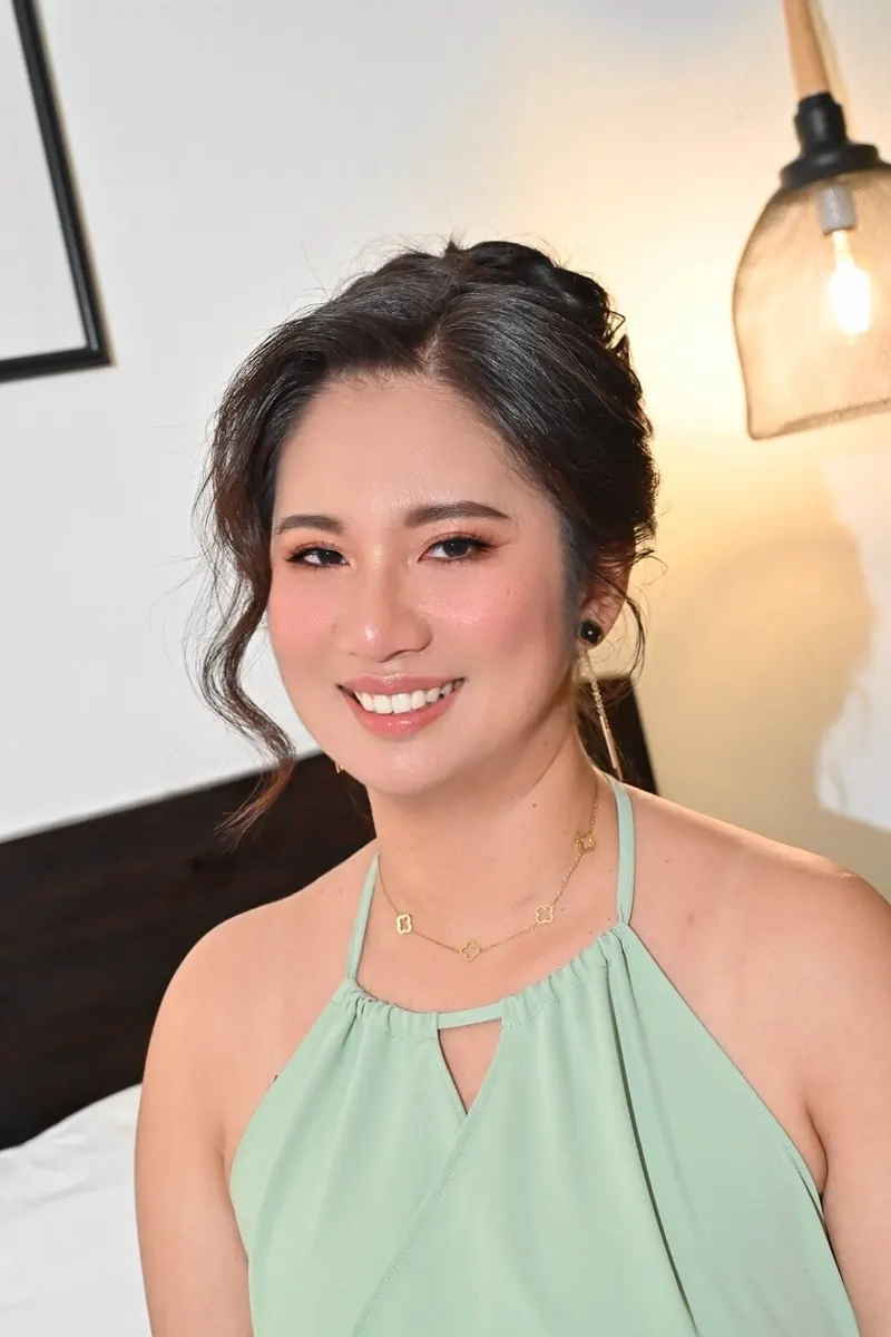 ชุดคล้องคอสีเขียวมิ้นต์ ผมเกล้าสูงปล่อยปอยข้างหน้า สวมสร้อยทองบาง ยิ้มกว้าง แต่งหน้าโทนพีชนู้ด โคมไฟห้อยด้านหลัง