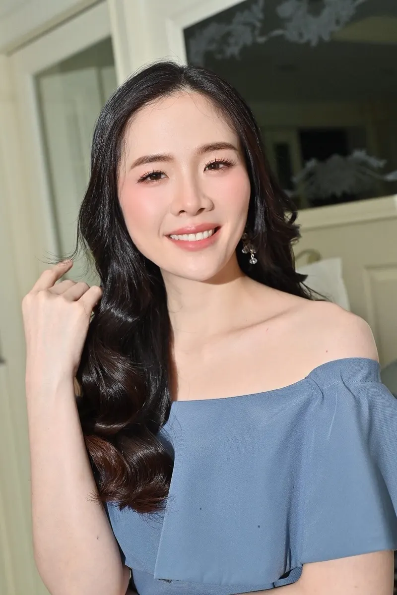 ผมดำลอนใหญ่ปล่อยด้านเดียว เสื้อเปิดไหล่สีฟ้าเทา สวมต่างหูห้อย แต่งหน้าโทนชมพูส้มสว่างใส ลิปสีชมพูนู้ด ฉากห้องสีขาว