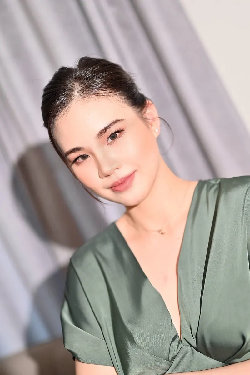 ผมดำรวบเก็บเรียบปล่อยปอยข้าง ชุดซาตินสีเขียวเทาคอวีทับหน้า ต่างหูไข่มุก แต่งหน้าบางเบา ลิปสีชมพูนู้ด ฉากผ้าม่านสีเทา