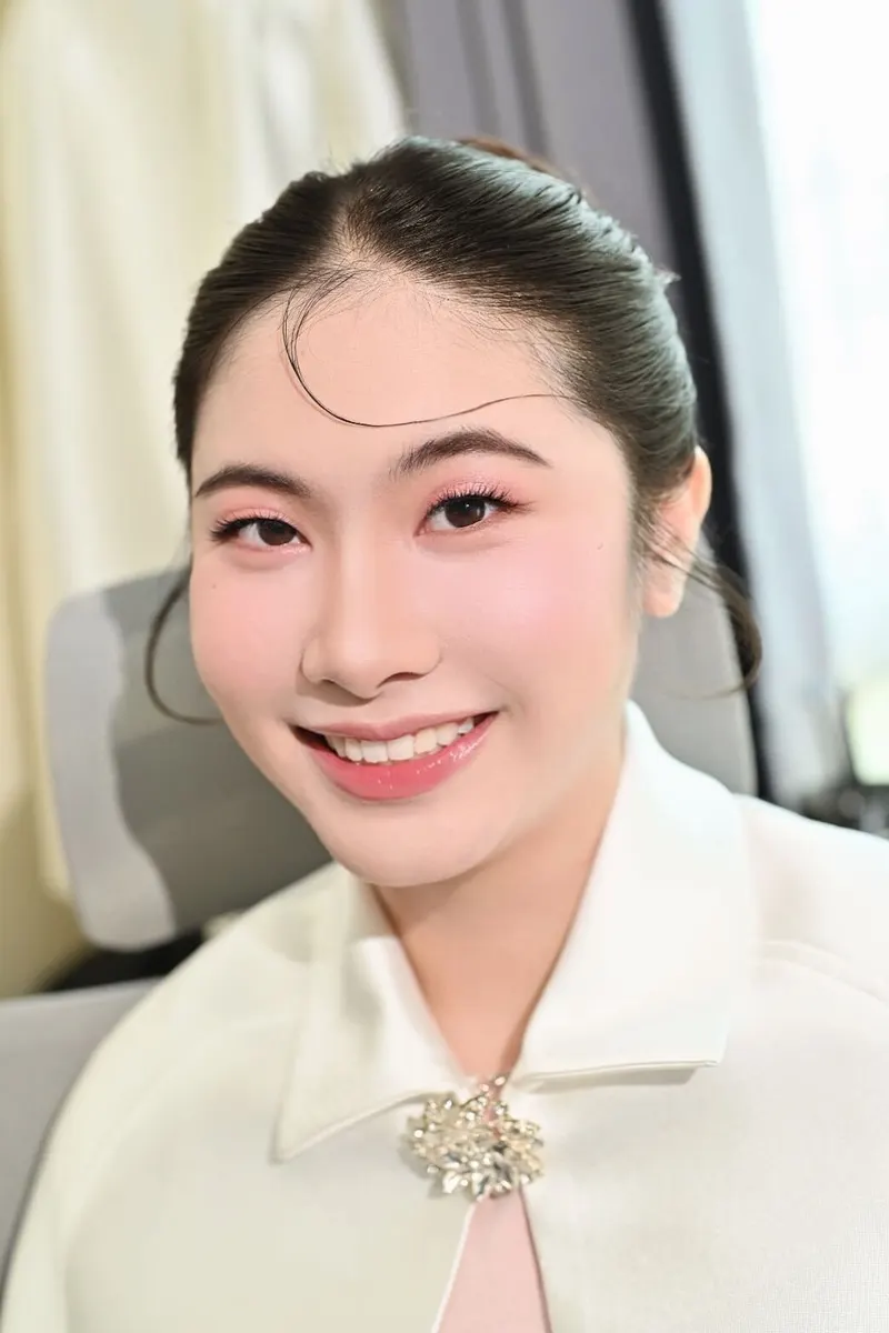 ผมดำรวบเก็บเรียบมีปอยสั้นข้างหน้า สวมเสื้อสีขาวมีเข็มกลัดเพชร แต่งตาชมพูแดงเน้นหางตา ปัดแก้มชมพูอ่อน ยิ้มเห็นฟัน