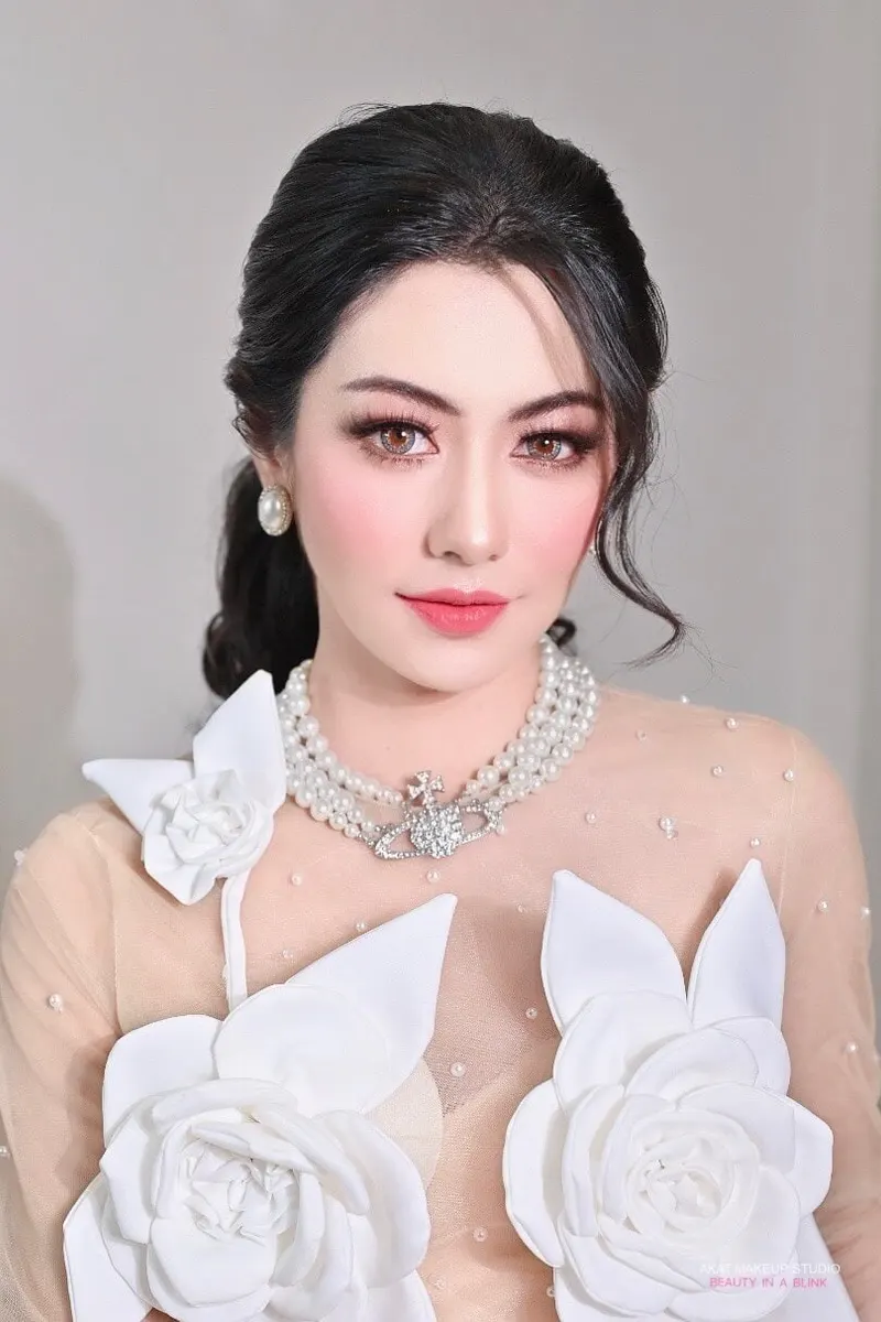 ผมดำเกล้าต่ำปล่อยลอนหางหมูข้างหู สวมชุดซีทรูปักไข่มุกประดับดอกกุหลาบผ้าสีขาว สร้อยไข่มุกหลายชั้น ปัดแก้มชมพูเด่น ลิปสีแดงชมพู