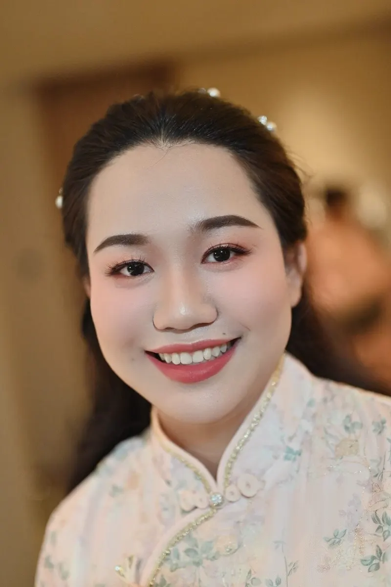 ผมดำรวบเก็บด้านหลังติดเข็มกลัดผมเล็ก ๆ สวมชุดจีนคอตั้งสีขาวลายดอกไม้ ยิ้มกว้าง แต่งหน้าโทนชมพูส้ม ลิปสีกุหลาบ ถ่ายภายในห้อง