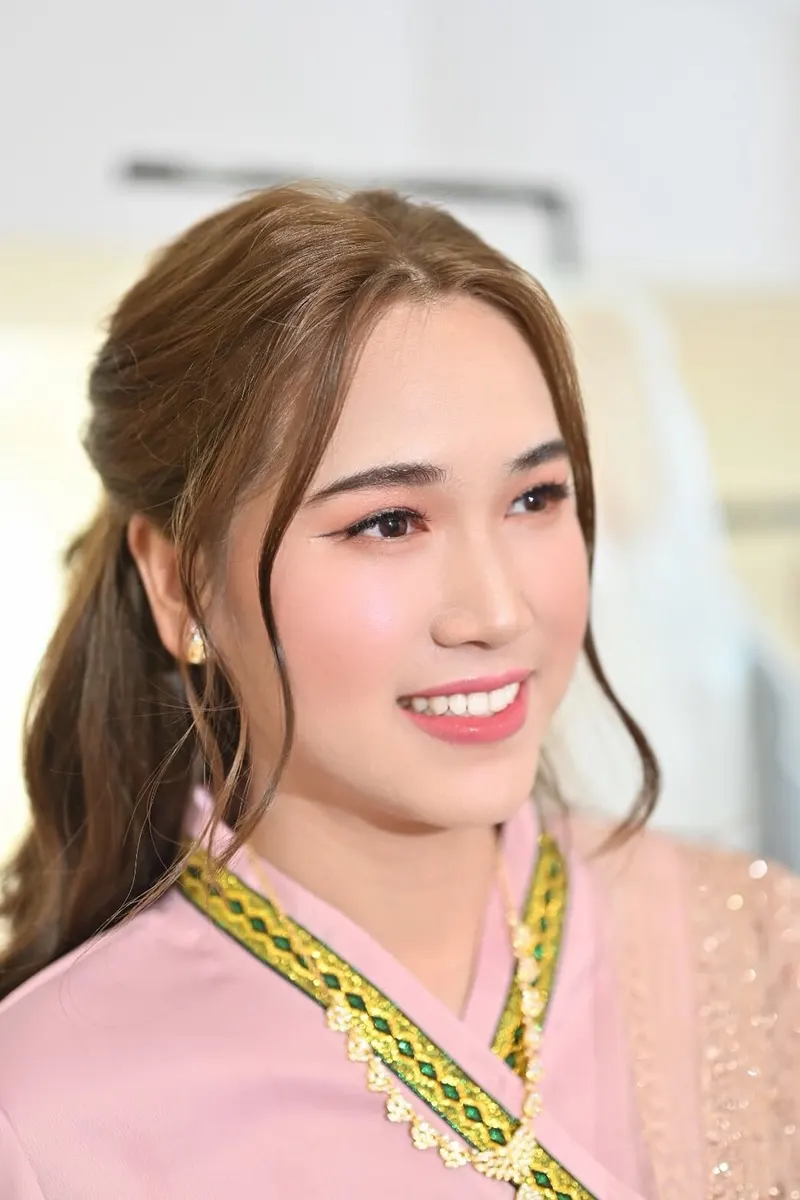 ผมน้ำตาลรวบครึ่งหัวปล่อยลอนข้างแก้ม ชุดไทยจีบสีชมพูคาดผ้าลายเขียวทอง ต่างหูมุก ยิ้มมองไปด้านข้าง แต่งตาชมพูพีช ลิปชมพูกลอส