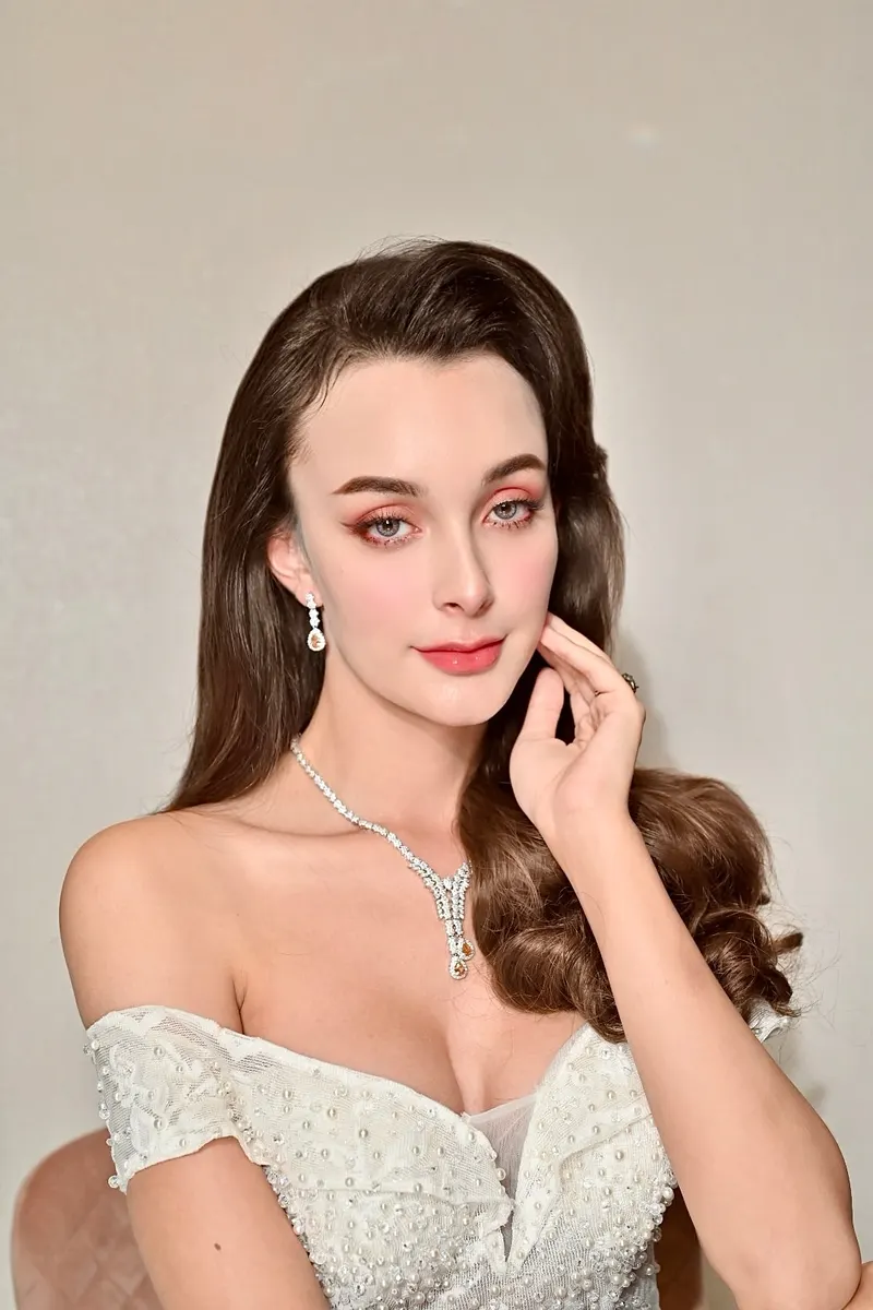 เจ้าสาวชุดวิวาห์เปิดไหล่ประดับไข่มุก ผมลอนใหญ่ปล่อยยาวด้านข้าง สวมสร้อยเพชร แต่งตาโทนพีชชมพู ลิปสีส้มอิฐ