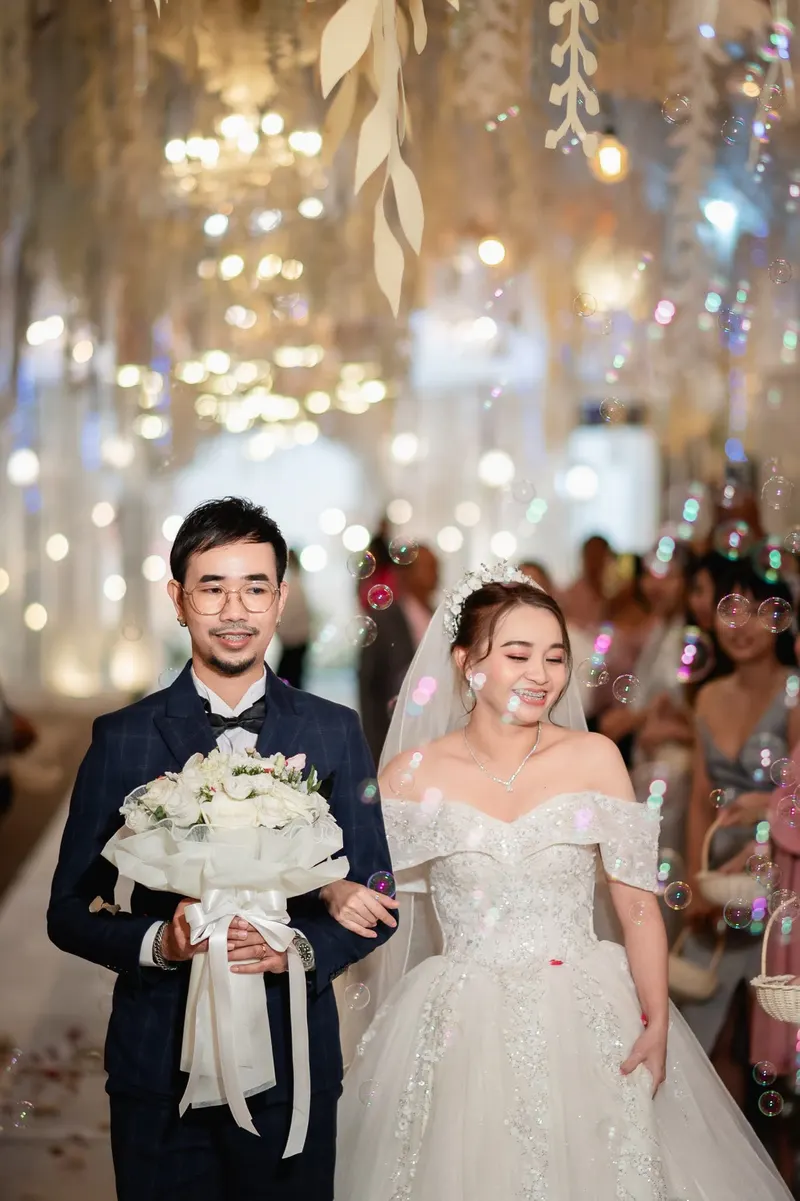คู่บ่าวสาวเดินเข้างานในโถงตกแต่งไฟระยิบ เจ้าสาวชุดบอลกาวน์เปิดไหล่สวมผ้าคลุมหน้า ถือช่อดอกไม้ขาว เจ้าบ่าวสูทกรมท่า