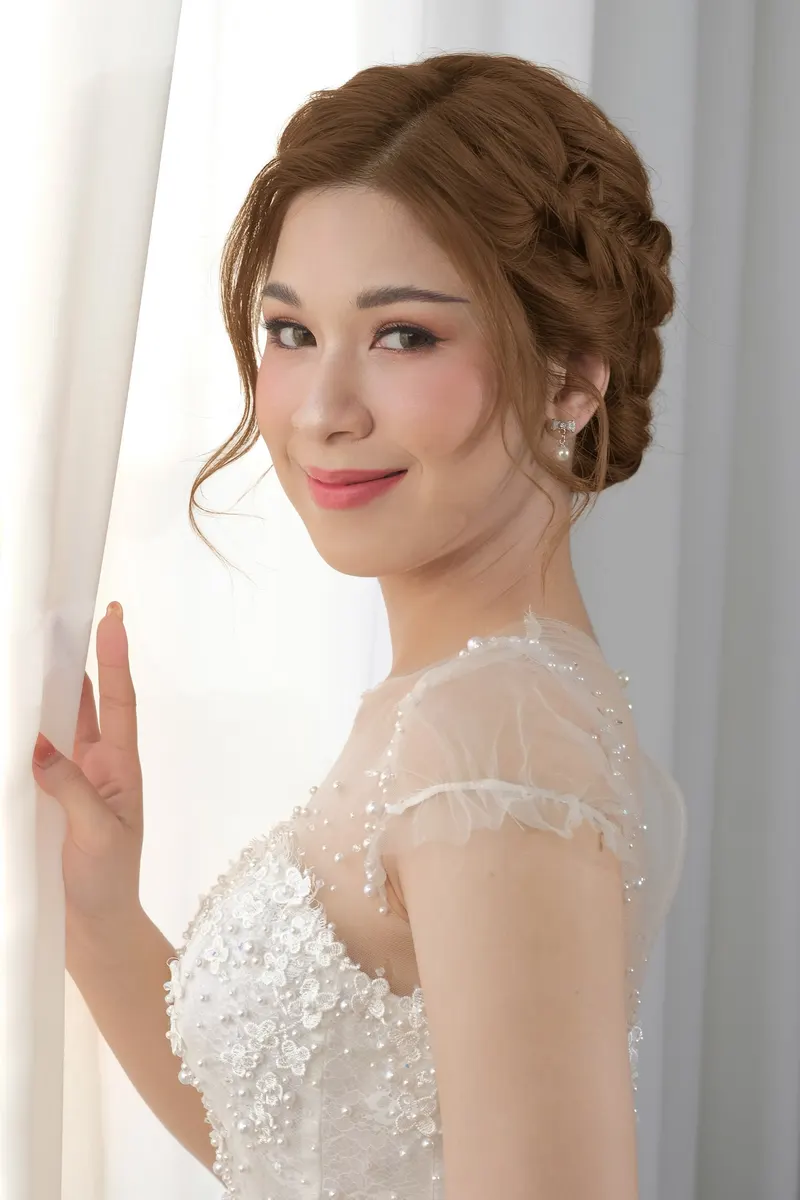 เจ้าสาวหันข้างริมผ้าม่านสีขาว ถักเปียเกล้าผมสีน้ำตาลทอง ชุดแขนสั้นตาข่ายปักไข่มุก ยิ้มบางๆ ลิปชมพูเข้ม