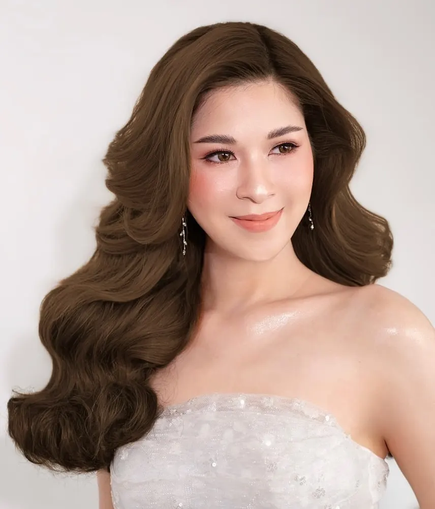 เจ้าสาวชุดเกาะอกลูกไม้ ผมลอนใหญ่สีน้ำตาลปล่อยด้านเดียว ต่างหูห้อยยาว ผิวฉ่ำ ปัดแก้มพีช ลิปส้มอ่อน ถ่ายบนพื้นหลังสีเทาอ่อน