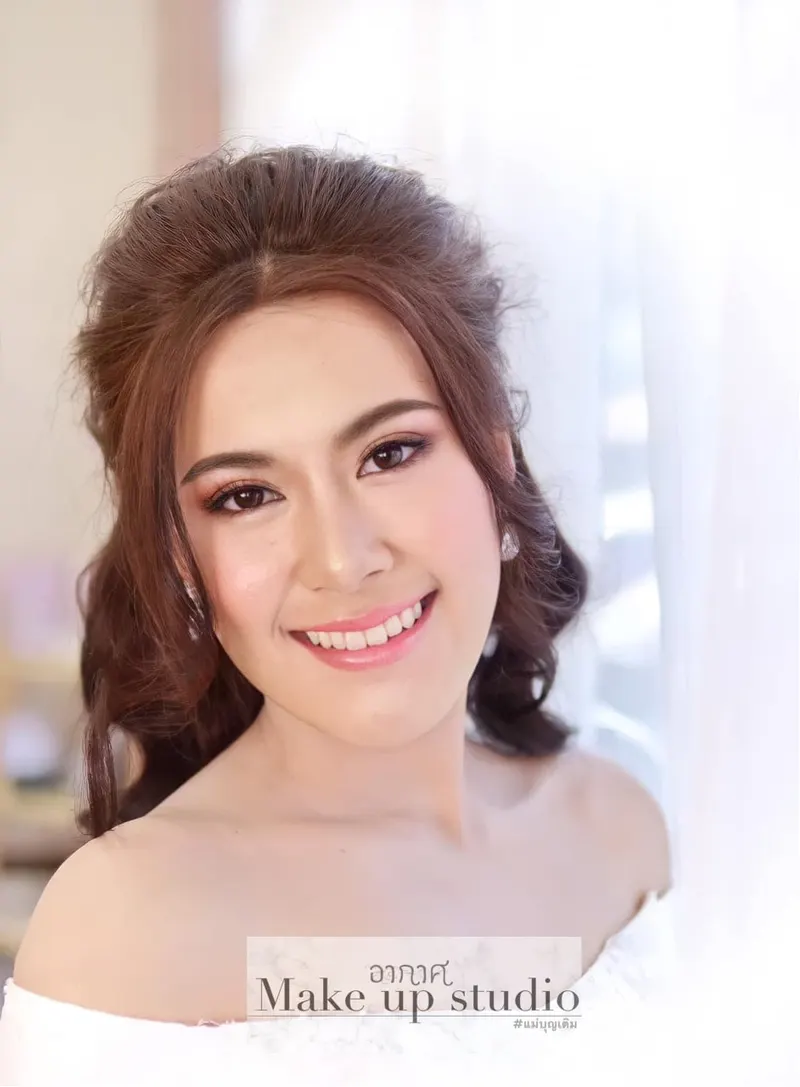 เจ้าสาวยิ้มสดใสหน้าผ้าม่านบางสีขาว ผมม้วนลอนเกล้าครึ่งหัวสีน้ำตาลแดง อายแชโดว์โทนม่วงน้ำตาล ลิปสีชมพูกลอส