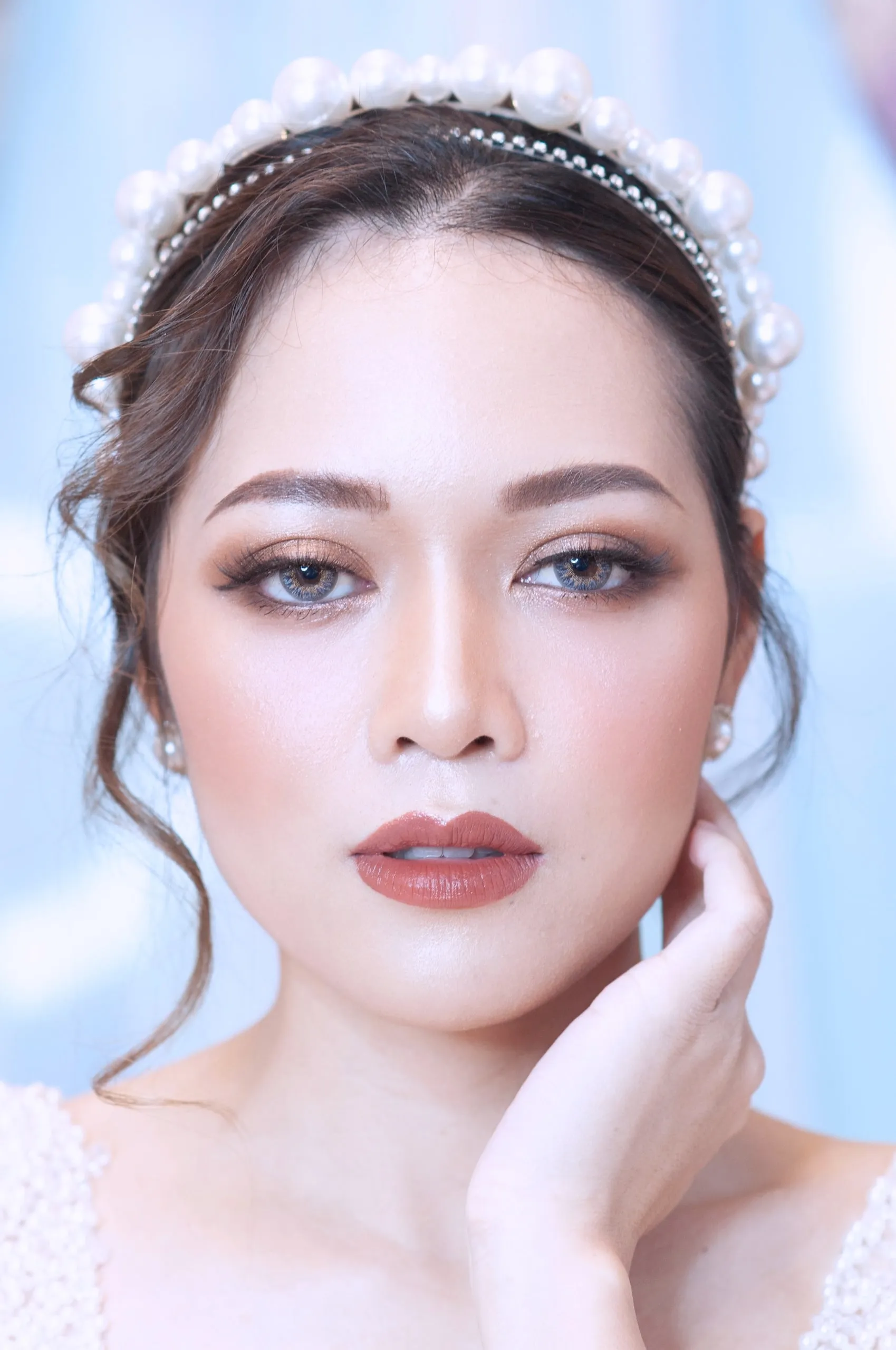 ภาพใกล้ใบหน้าเจ้าสาวสวมที่คาดผมไข่มุกเม็ดโต แต่งตาสโมกกี้น้ำตาล ลิปสีน้ำตาลแดง ผิวหน้าเรียบมีประกาย เอามือแตะข้างคอ