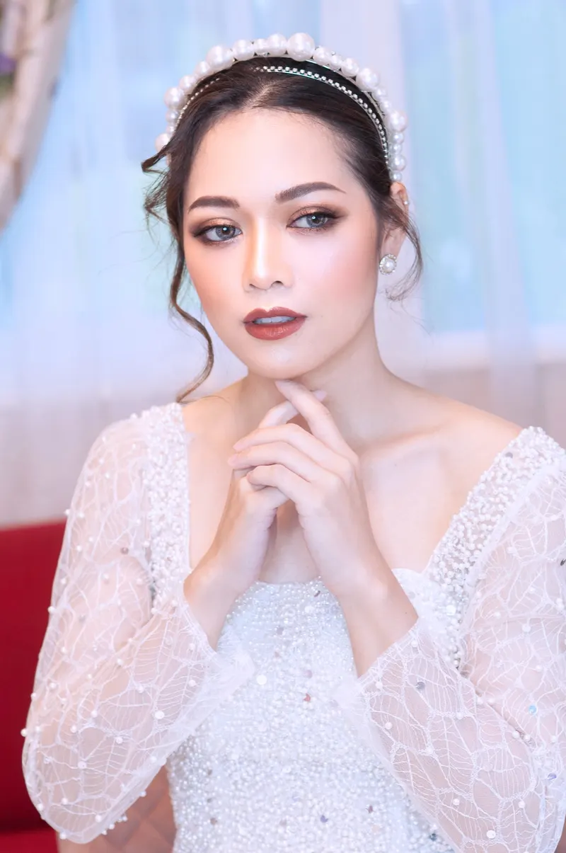 เจ้าสาวชุดลูกไม้แขนยาวประดับไข่มุก ที่คาดผมไข่มุก ยกสองมือประสานใต้คาง แต่งตาน้ำตาลเข้ม ลิปสีไวน์
