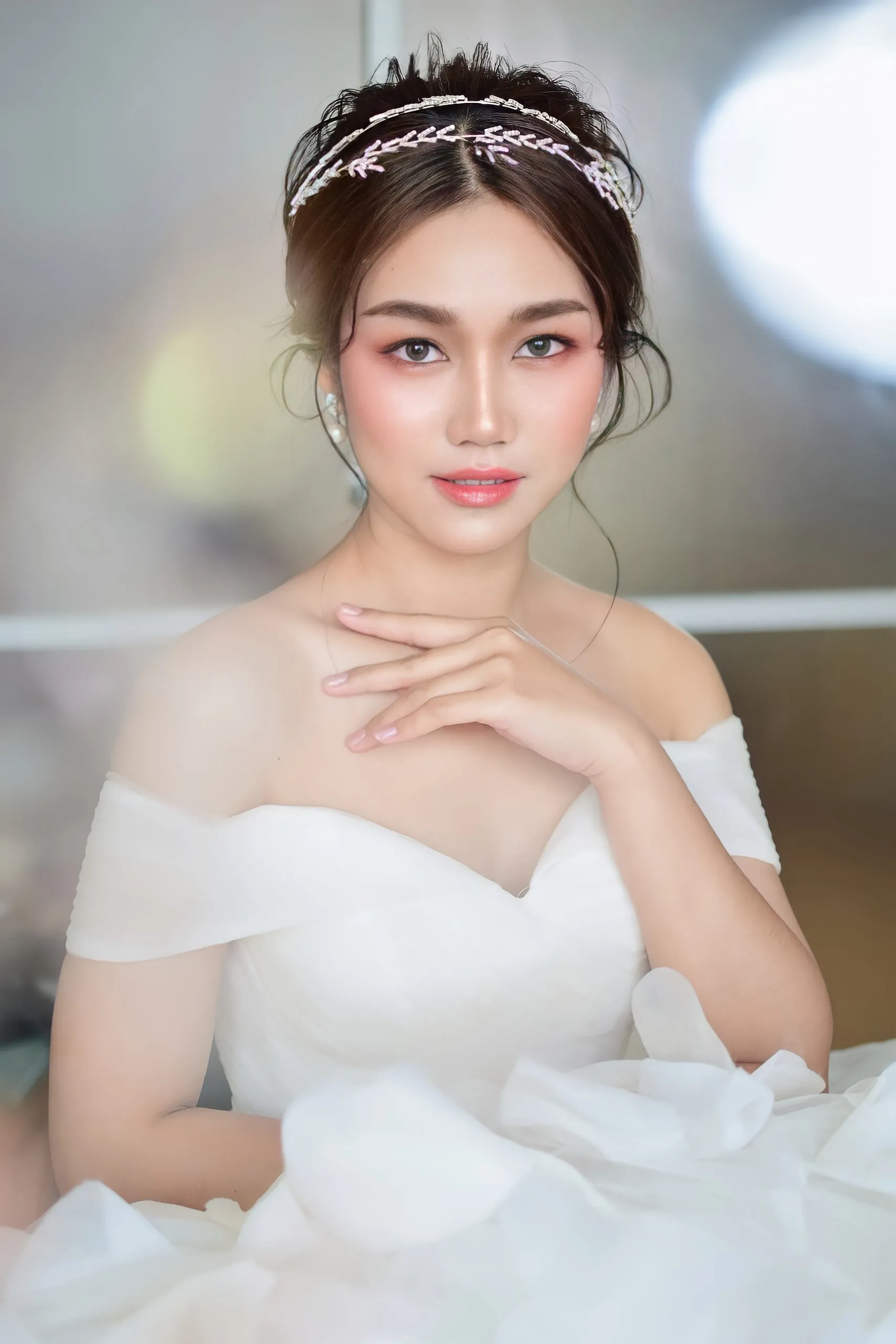 เจ้าสาวสวมที่คาดผมคริสตัลทรงใบไม้ชุดเปิดไหล่ผ้าฟุ้ง ผมเกล้าหลวม วางมือแนบอก แก้มชมพูส้มและลิปชมพูพีช ถ่ายในแสงนุ่มหน้าฉากเบลอ