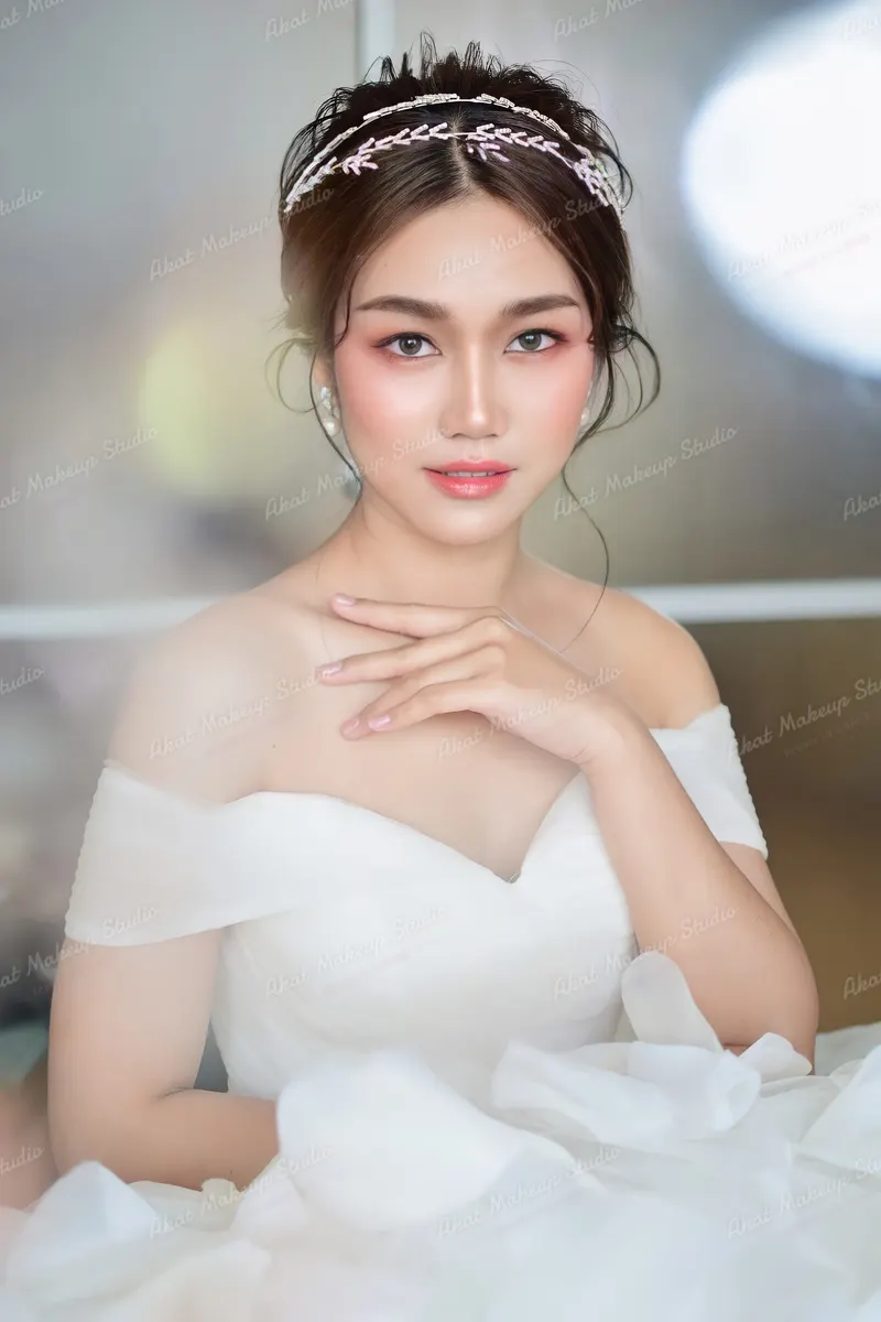เจ้าสาวชุดเปิดไหล่ผ้าฟุ้ง สวมที่คาดผมคริสตัลทรงใบไม้ ผมเกล้าสูงหลวมๆ ปัดแก้มชมพูส้มเด่น ลิปชมพูพีช