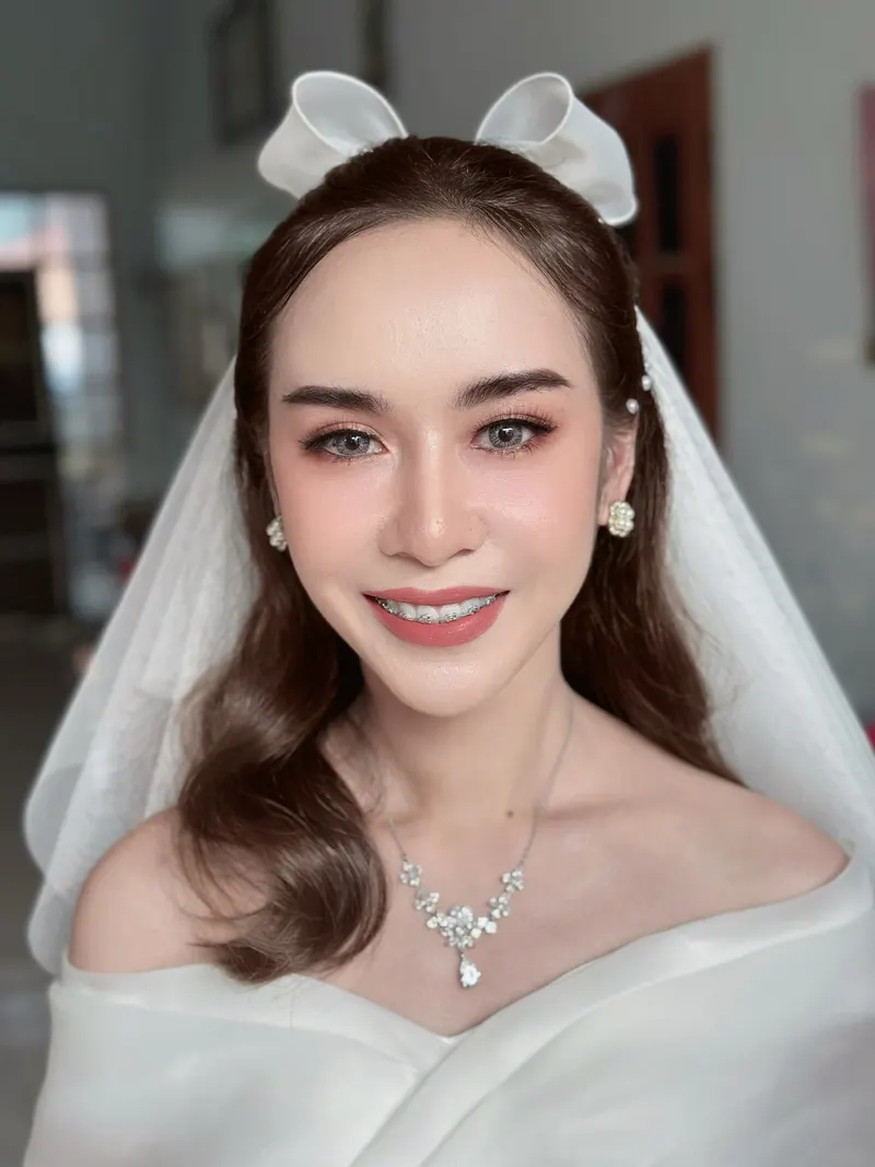 เจ้าสาวสวมผ้าคลุมหน้ากับโบว์ผ้าตาข่ายเหนือศีรษะ ผมยาวสีน้ำตาลลอนอ่อน สร้อยคอเพชรช่อดอกไม้ แต่งผิวสว่างใส ลิปสีนู้ดชมพู