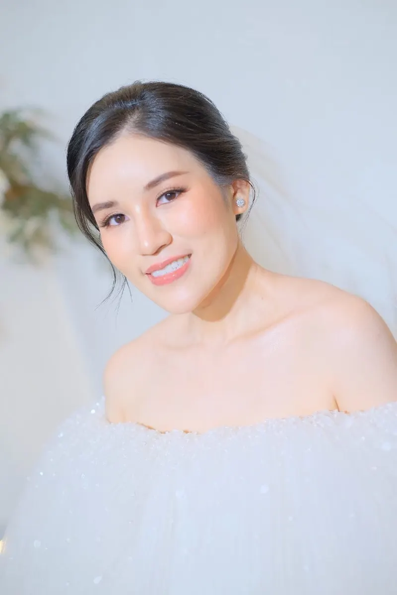 เจ้าสาวไหล่เปลือยชุดทูลเฟ้อฟู่สีขาว ผมรวบตึงคลุมผ้าคลุมหน้าบางๆ ยิ้มเอียงศีรษะ แต่งหน้าโทนพีชธรรมชาติ แสงสว่างนุ่ม