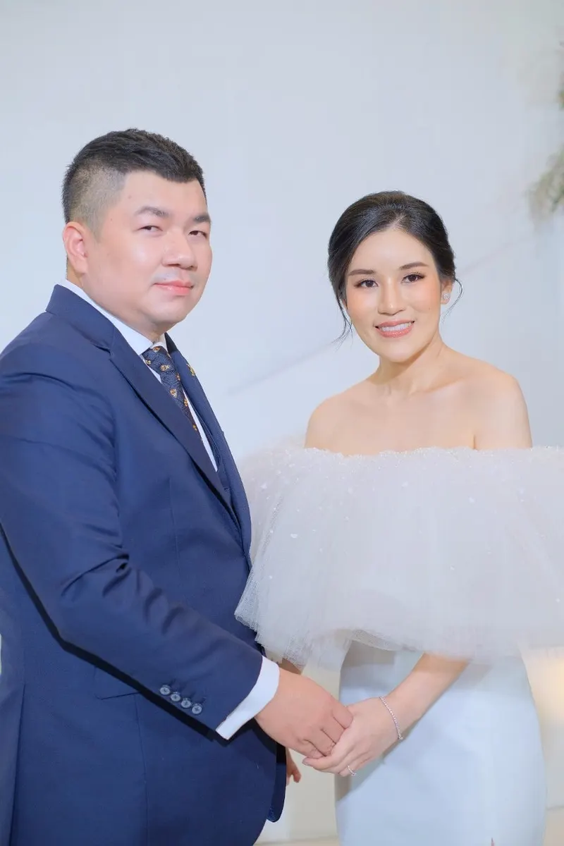 คู่บ่าวสาวยืนจับมือกันในสตูดิโอฉากขาว เจ้าสาวชุดทูลเปิดไหล่ ผมเกล้าเก็บ เจ้าบ่าวสูทกรมท่า แต่งหน้าโทนนุ่มบางเบา