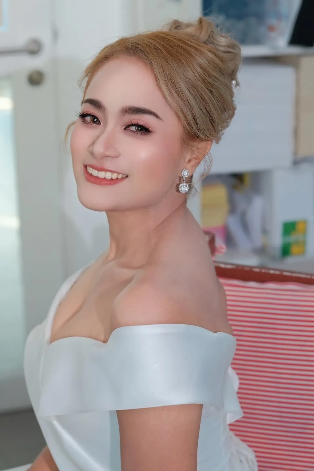 เจ้าสาวผมสีบลอนด์เกล้ามวยสูง สวมชุดซาตินเปิดไหล่และต่างหูเพชรเหลี่ยม หันข้างยิ้มกว้าง แต่งตาน้ำตาลอ่อน ลิปสีส้มพีช อยู่ในห้องที่มีแสงจากหน้าต่าง