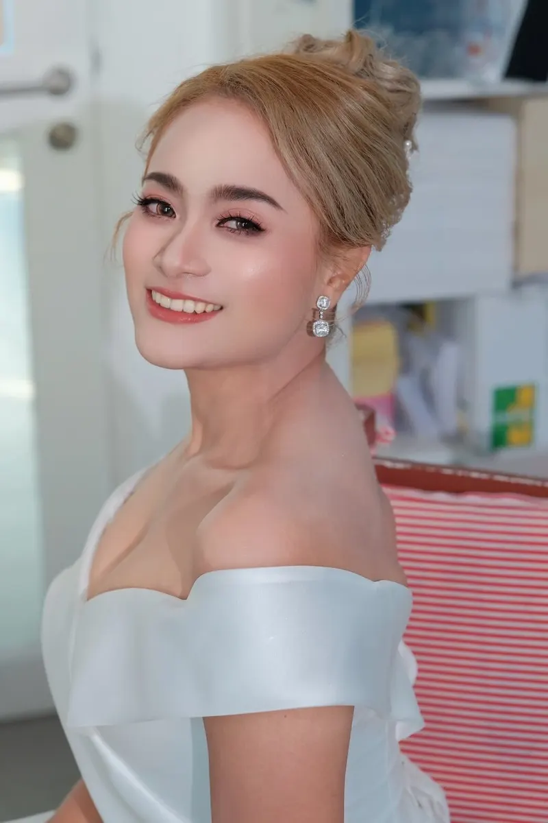 เจ้าสาวหันข้างยิ้มมองกล้อง ผมสีบลอนด์ทองเกล้ามวยสูง ต่างหูเพชรเหลี่ยม ชุดซาตินเปิดไหล่ แต่งตาน้ำตาลอ่อน ลิปสีส้มพีช