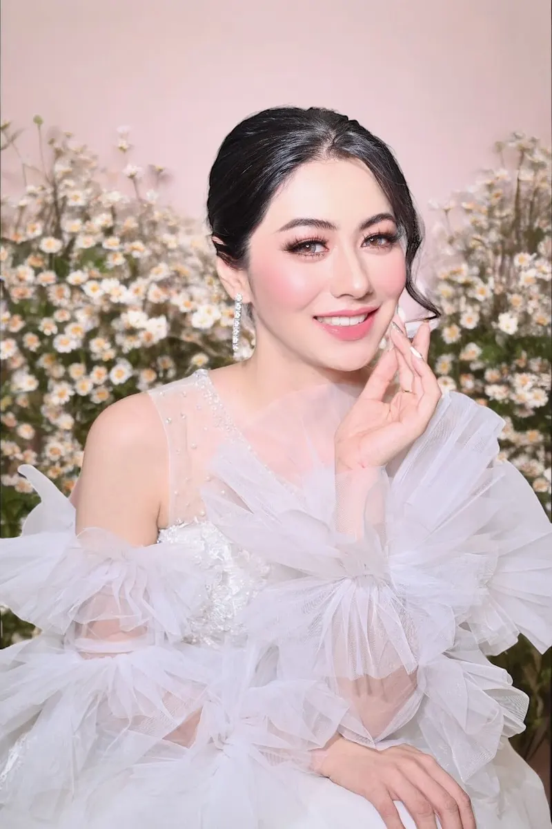 เจ้าสาวนั่งหน้าพุ่มดอกเดซี่สีขาว ชุดทูลระบายหลายชั้นแขนพองสีขาว ผมดำรวบข้างลอนปล่อย ต่างหูระย้า ปัดแก้มชมพู ลิปสีชมพูกลอส