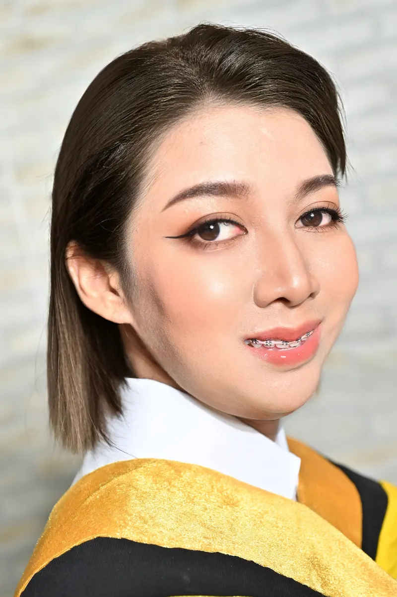 แต่งหน้ารับปริญญา ตาหางเฉียงเขียนอายไลเนอร์ปลายเรียว ลิปสีส้มพีช ผมสั้นประบ่าเรียบตรง สวมครุยดำคาดผ้าสีทอง ฉากผนังอิฐ