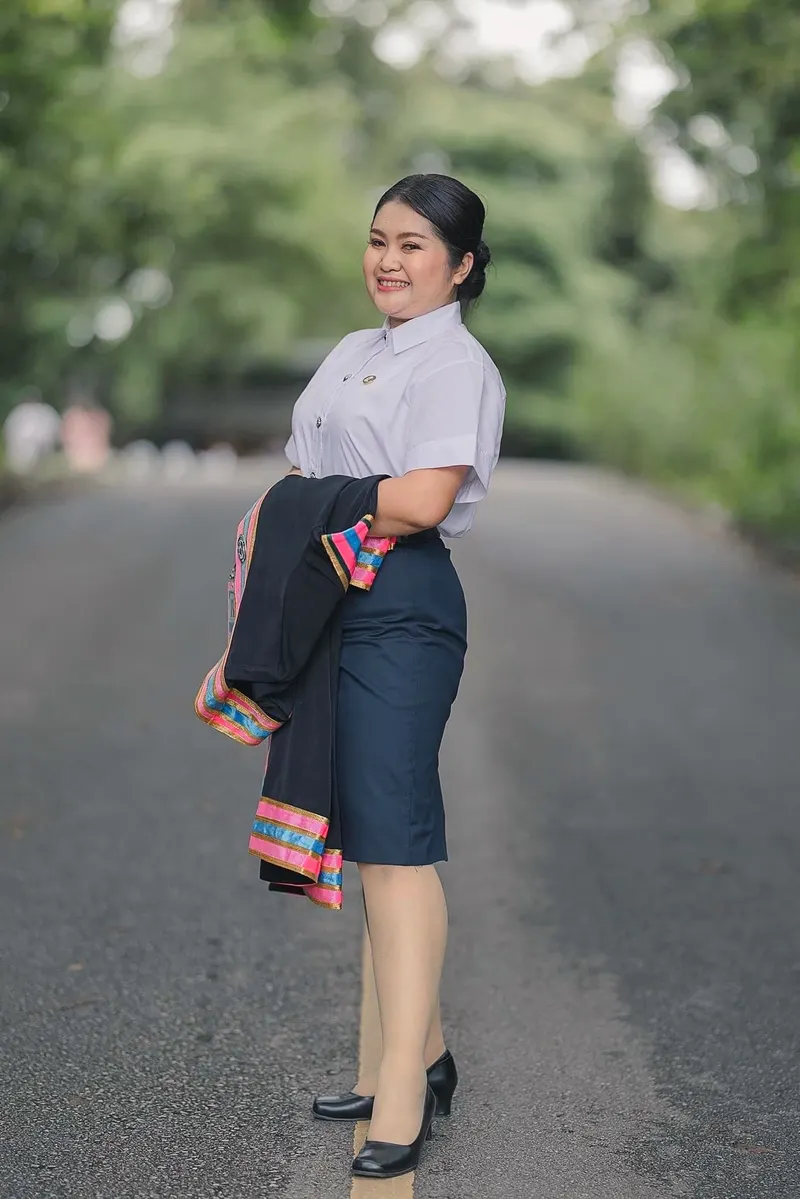ภาพเต็มตัวบัณฑิตหญิงยืนบนถนนในสวน ผมมวยต่ำ เสื้อนักศึกษาสีขาวกับกระโปรงกรมท่า พาดครุยดำขอบชมพูไว้บนแขน