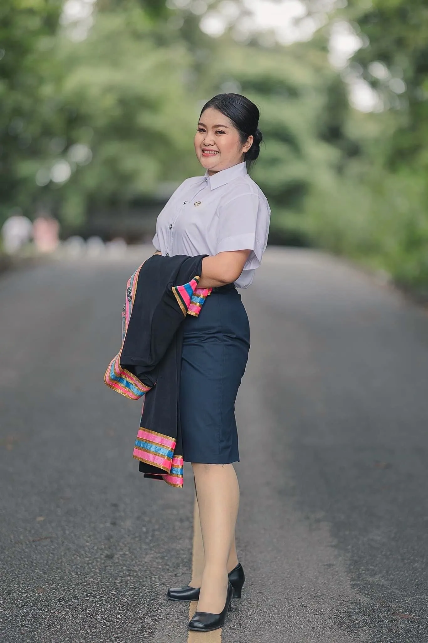 บัณฑิตหญิงยืนบนถนนในสวน ผมมวยต่ำ เสื้อนักศึกษาสีขาวกับกระโปรงกรมท่า พาดครุยดำขอบชมพูไว้บนแขน