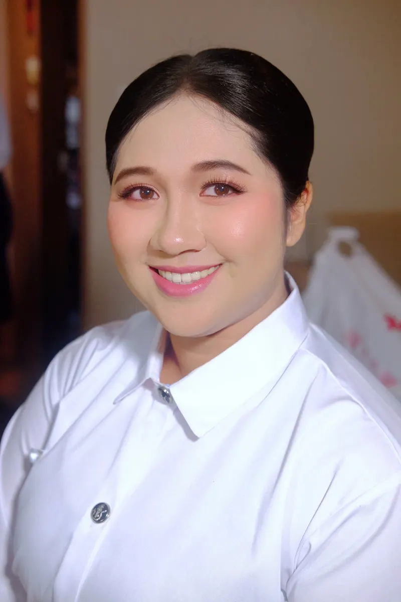 ผมรวบตึงเก็บเรียบหลังใบหู แต่งตาโทนส้มพีช ปัดแก้มชมพู ลิปชมพูกลอส ยิ้มกว้างในเสื้อนักศึกษาสีขาวติดกระดุมโลหะ