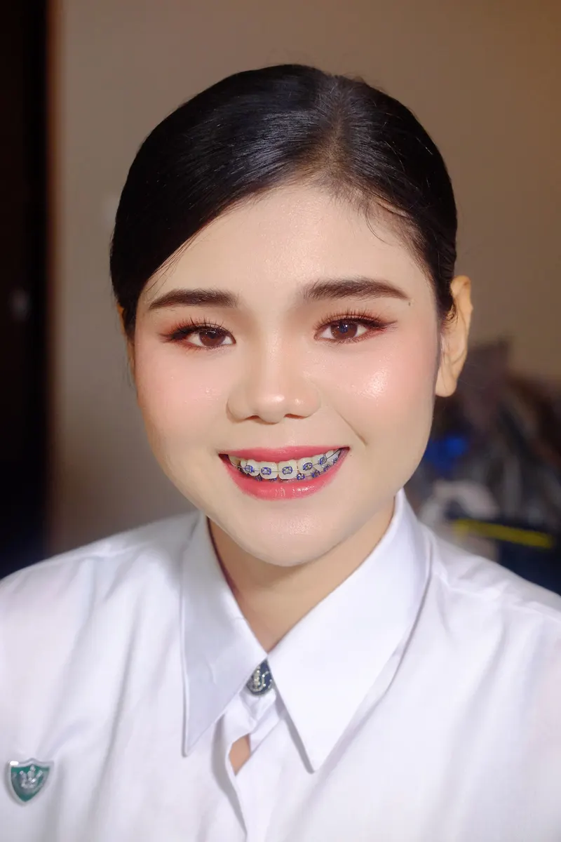 ผมดำรวบตึงเรียบ แต่งตาส้มทองเน้นหางตา ปัดแก้มสีพีชสดใส ลิปสีส้มแดง ยิ้มกว้างในชุดนักศึกษาสีขาวติดโลโก้