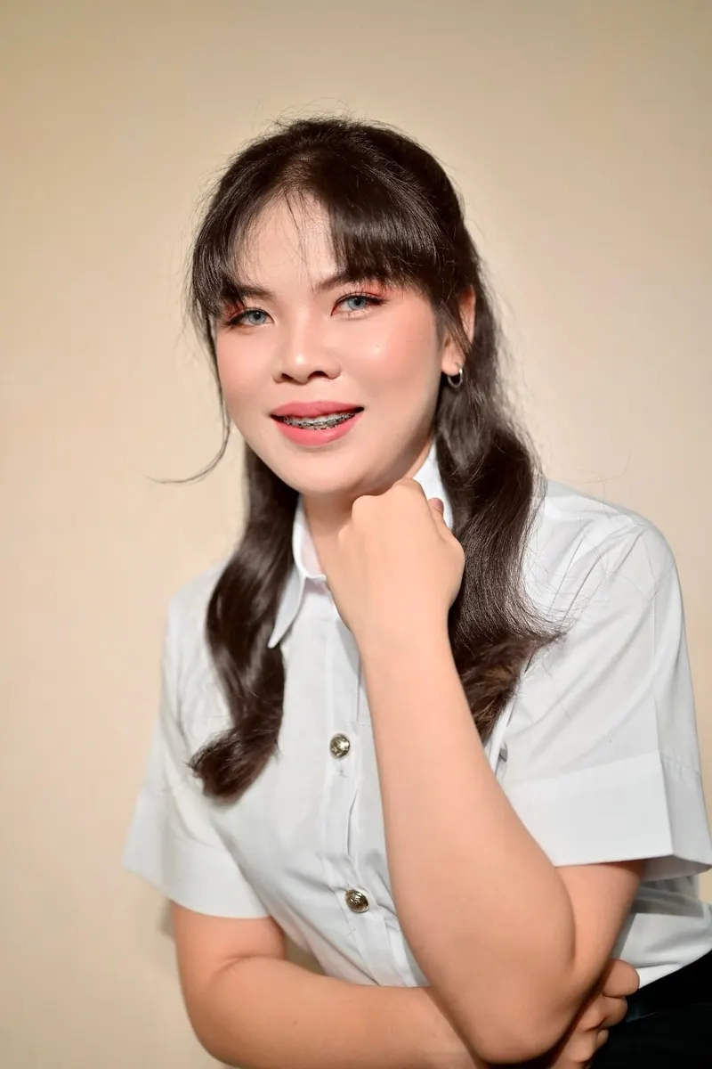 ผมม้าหน้าปัดบางผมยาวลอนข้างอกสองข้าง แต่งตาชมพูแดงและปัดแก้มสด ลิปสีแดงคอรัล ท่าเอามือแตะคางในเสื้อนักศึกษาสีขาว