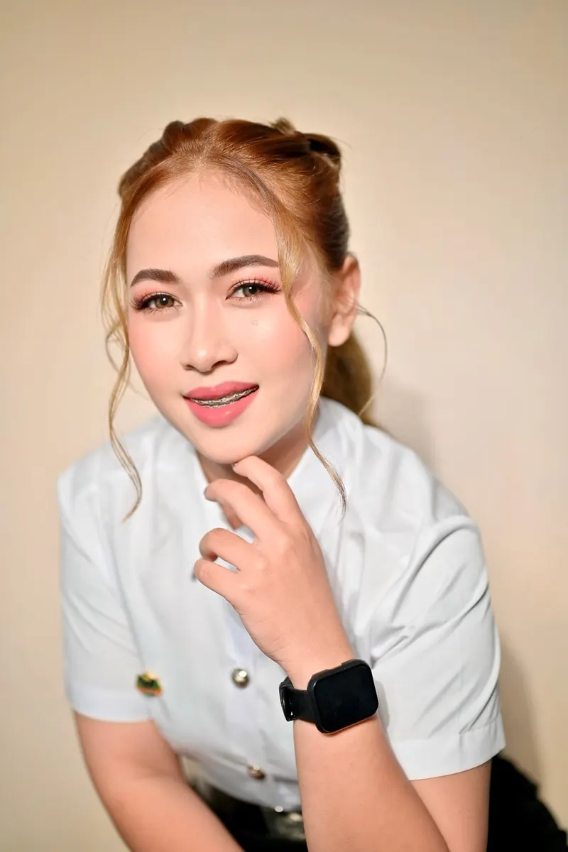 ผมสีน้ำตาลอ่อนทองรวบมวยสองข้างปล่อยปอยหน้า แต่งตาโทนทองชมพู ลิปสีชมพูเข้ม ยกมือแตะคาง สวมนาฬิกาข้อมือสีดำ