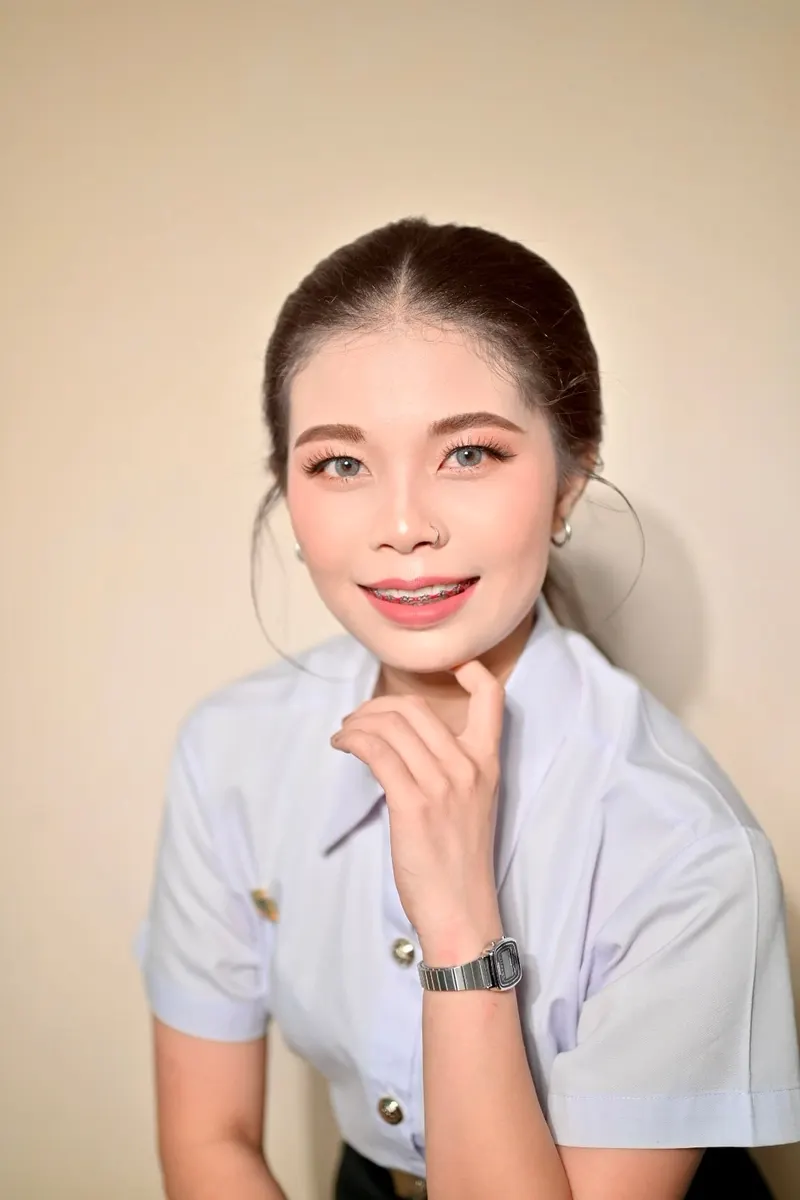 ผมเกล้ามวยต่ำเรียบ แต่งตาโทนส้มทอง ปัดแก้มชมพูพีช ลิปสีนู้ดชมพู เอามือแตะคาง เสื้อนักศึกษาสีขาวอมเทา พื้นหลังสีครีม