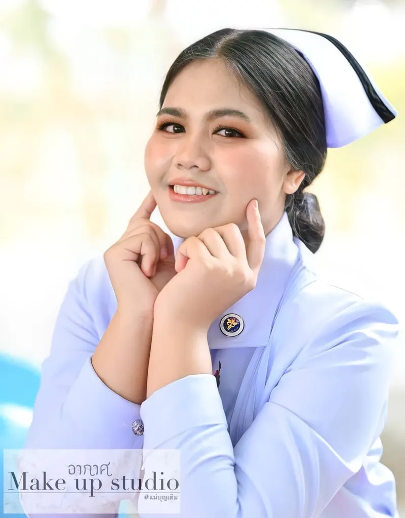 นักศึกษาพยาบาลสวมหมวกพยาบาลสีขาวขอบดำ ผมรวบต่ำ ยิ้มกว้างเอามือประสานแตะแก้ม แต่งตาโทนน้ำตาลทอง ลิปสีส้มพีช