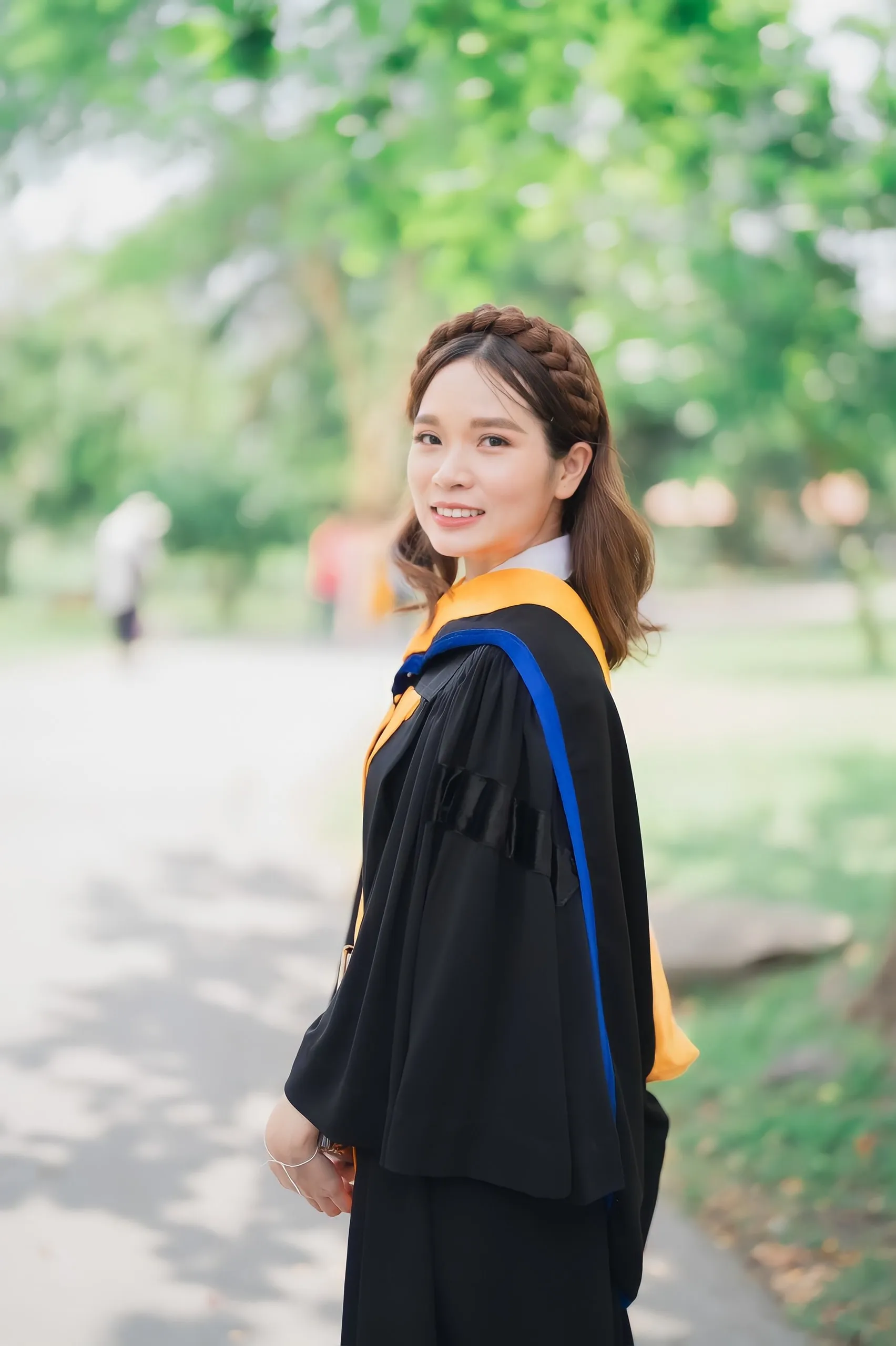 บัณฑิตสวมครุยดำคาดผ้าสีส้มและน้ำเงิน ผมถักเปียรอบศีรษะปล่อยปลายลอน หันข้างยิ้มกลางต้นไม้เขียว ในแสงกลางวัน